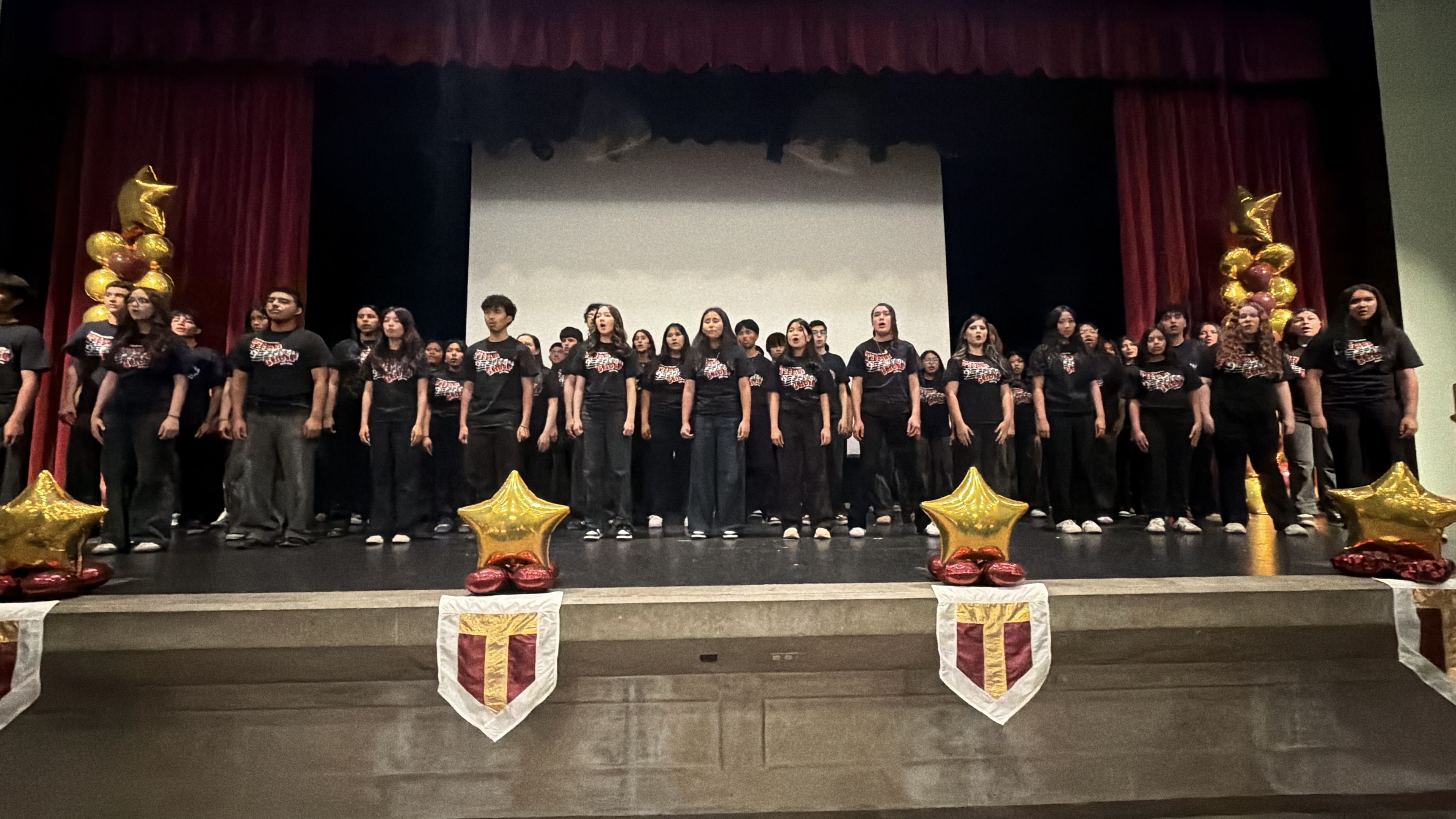 A large group of people, likely a choir or performance group, standing on a stage with red curtains and decorative elements in the background.