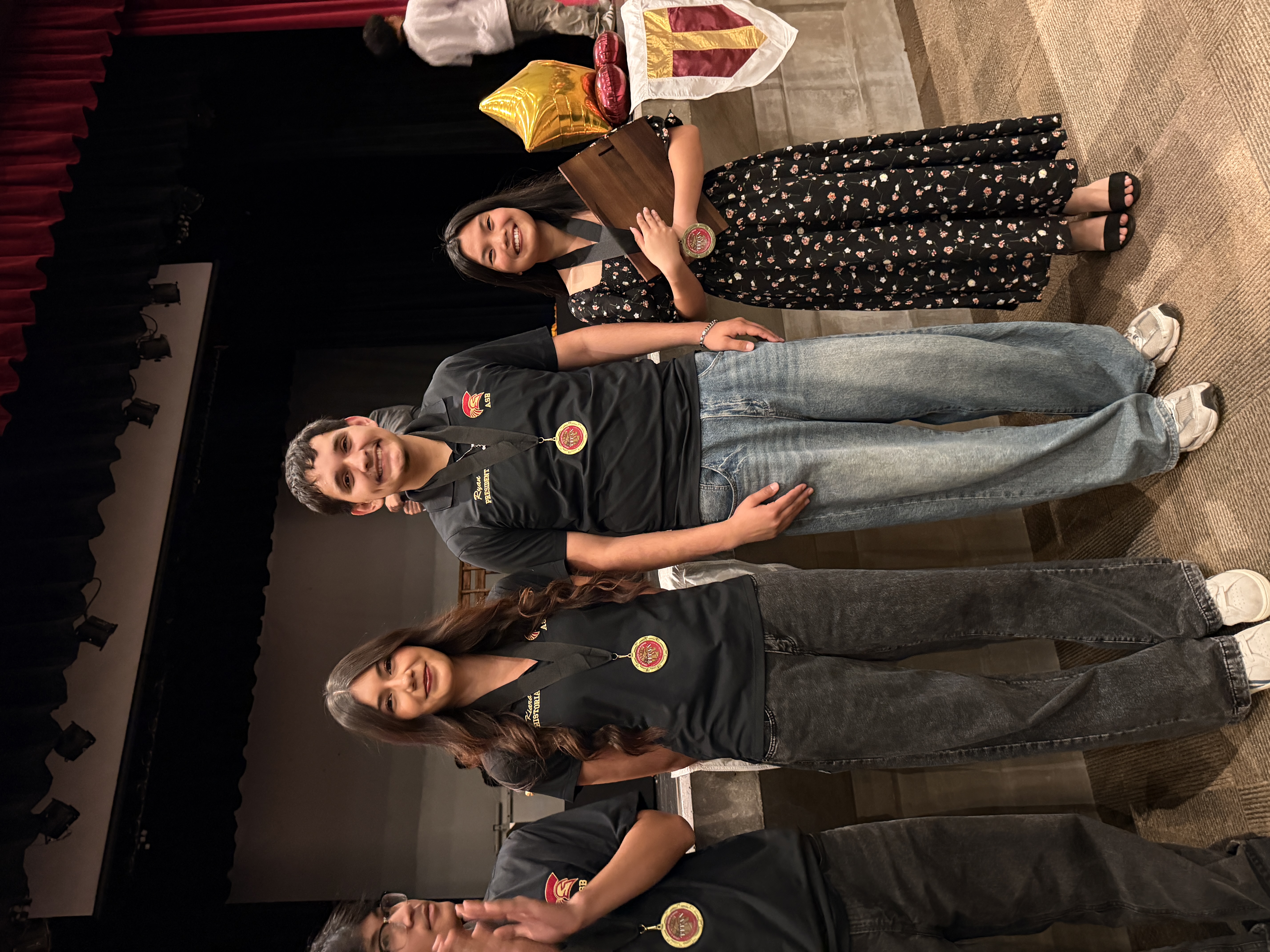 The image shows a group of people standing on a stage, dressed in casual attire and posing for a photograph. The background appears to be a theater or performance venue, with a red curtain visible behind them.