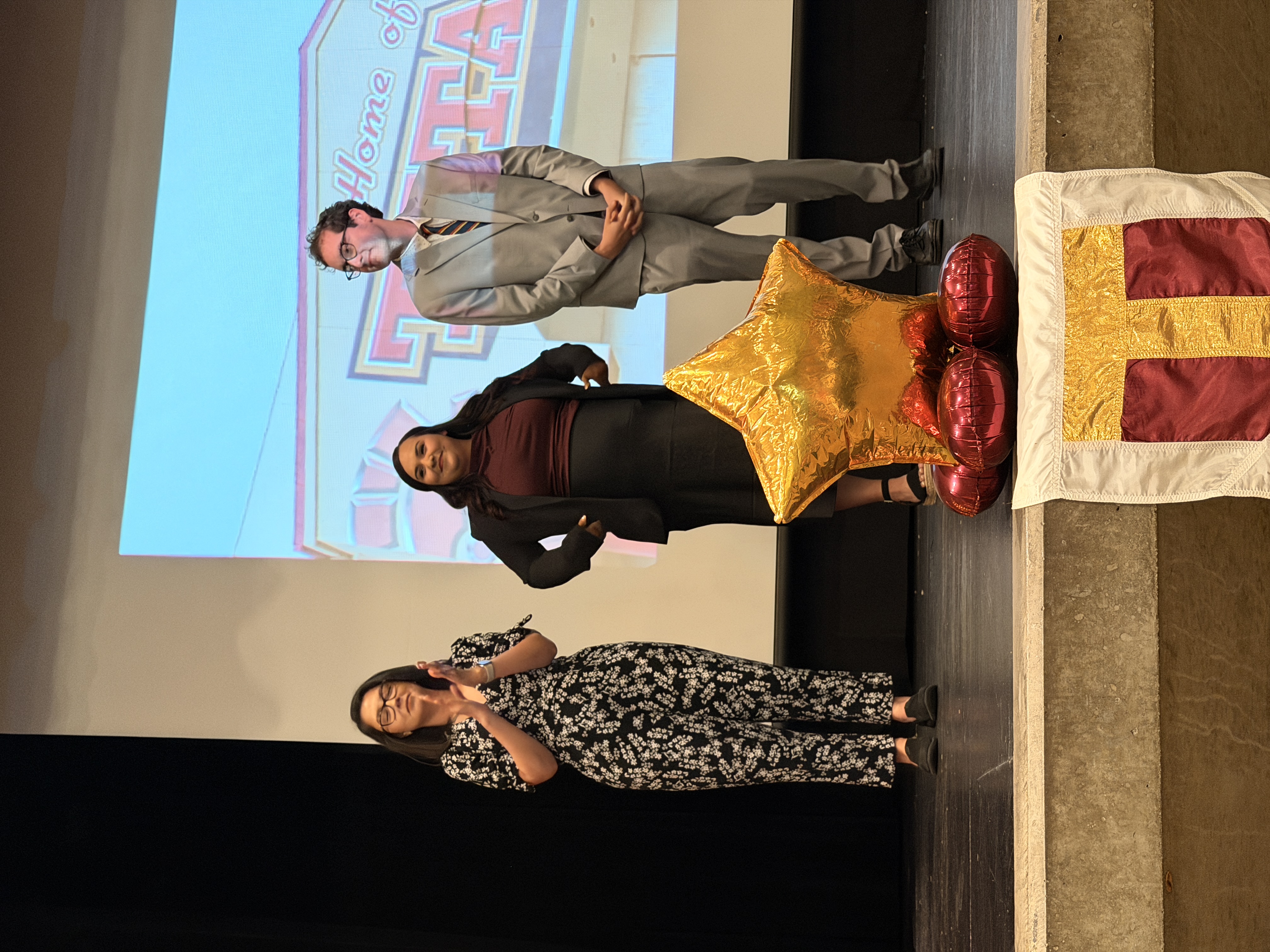Three individuals, two women and one man, stand on a stage with a large screen displaying a colorful background. In the foreground, a large golden star-shaped object is prominently displayed.