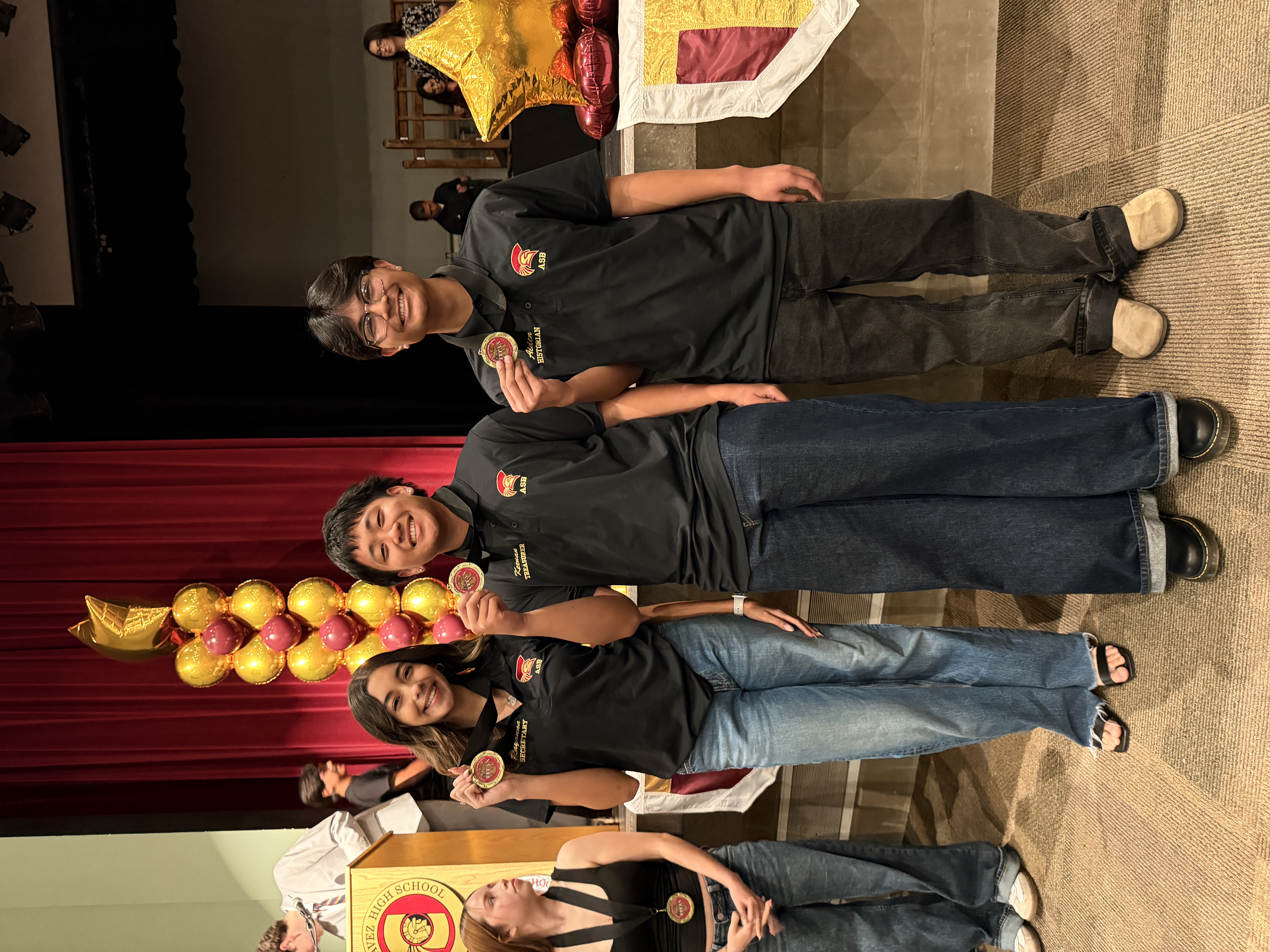 The image shows a group of people in casual attire, including black shirts with a logo, standing in front of a red curtain backdrop with a decorative display of golden objects.