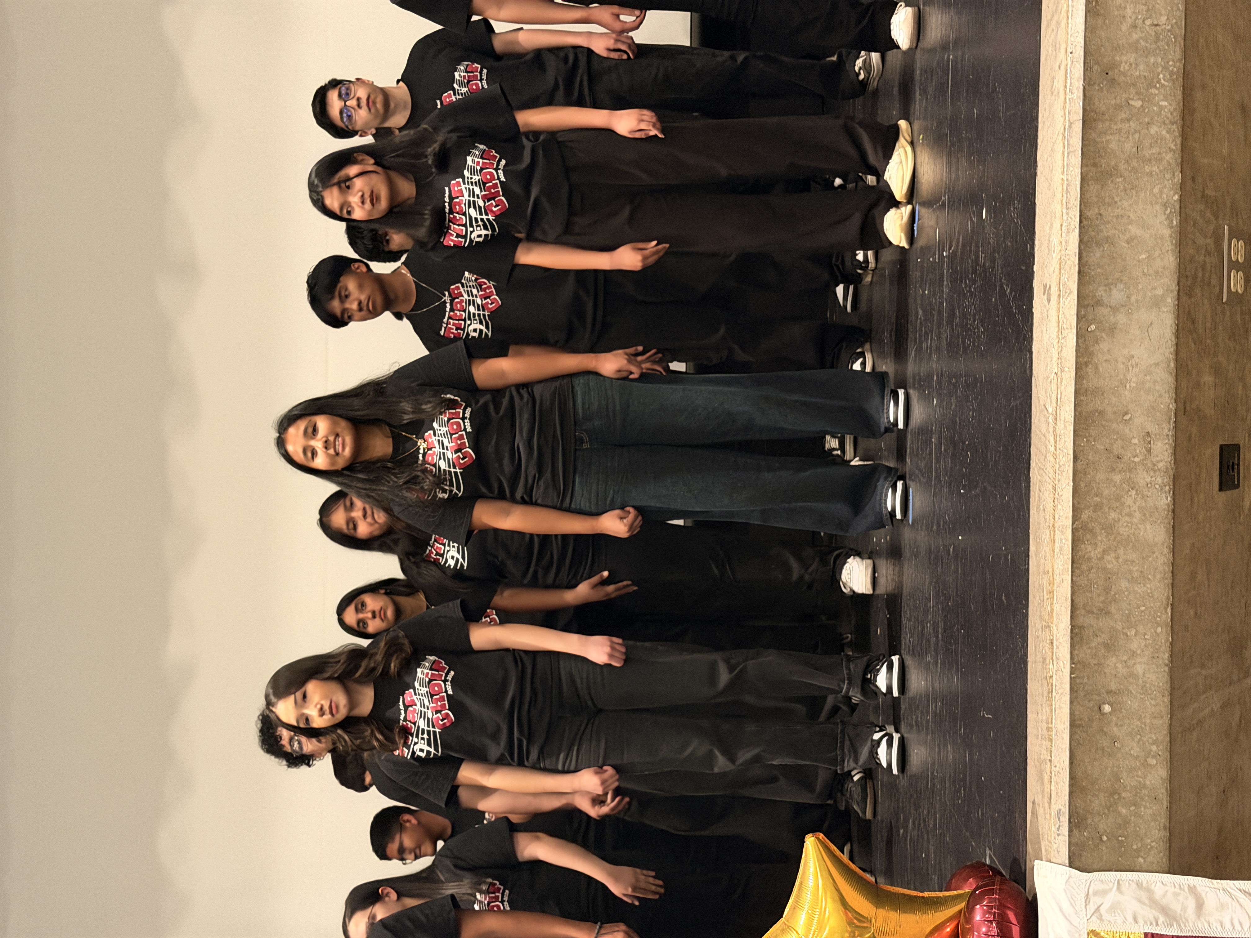 A group of young people, likely a dance or performance troupe, standing on a stage in coordinated black outfits with text on their shirts, against a backdrop of a yellow prop or set piece.