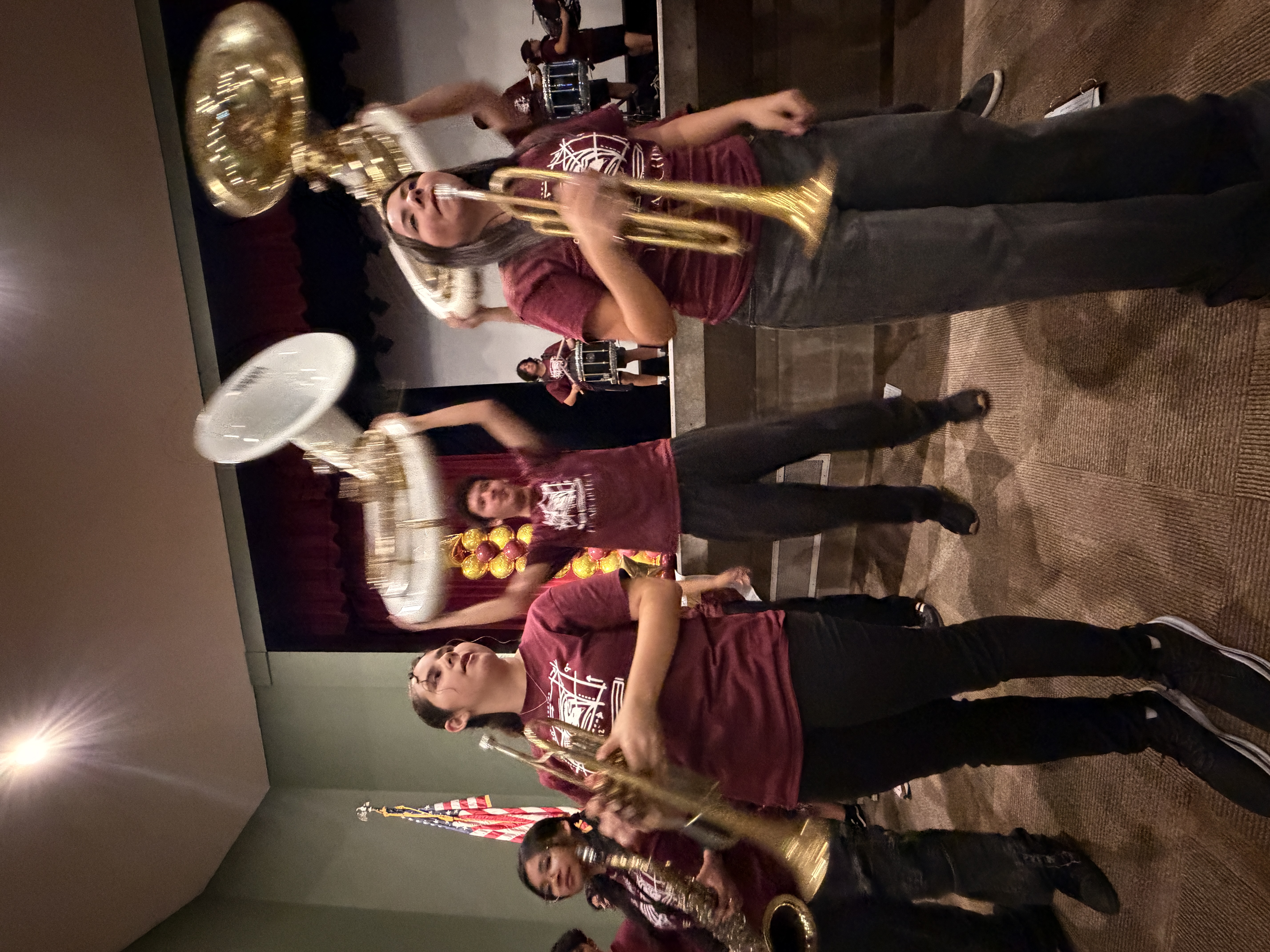A group of people wearing maroon shirts and holding various musical instruments, including trumpets and drums, appear to be performing or rehearsing in a room with a stage and lighting.