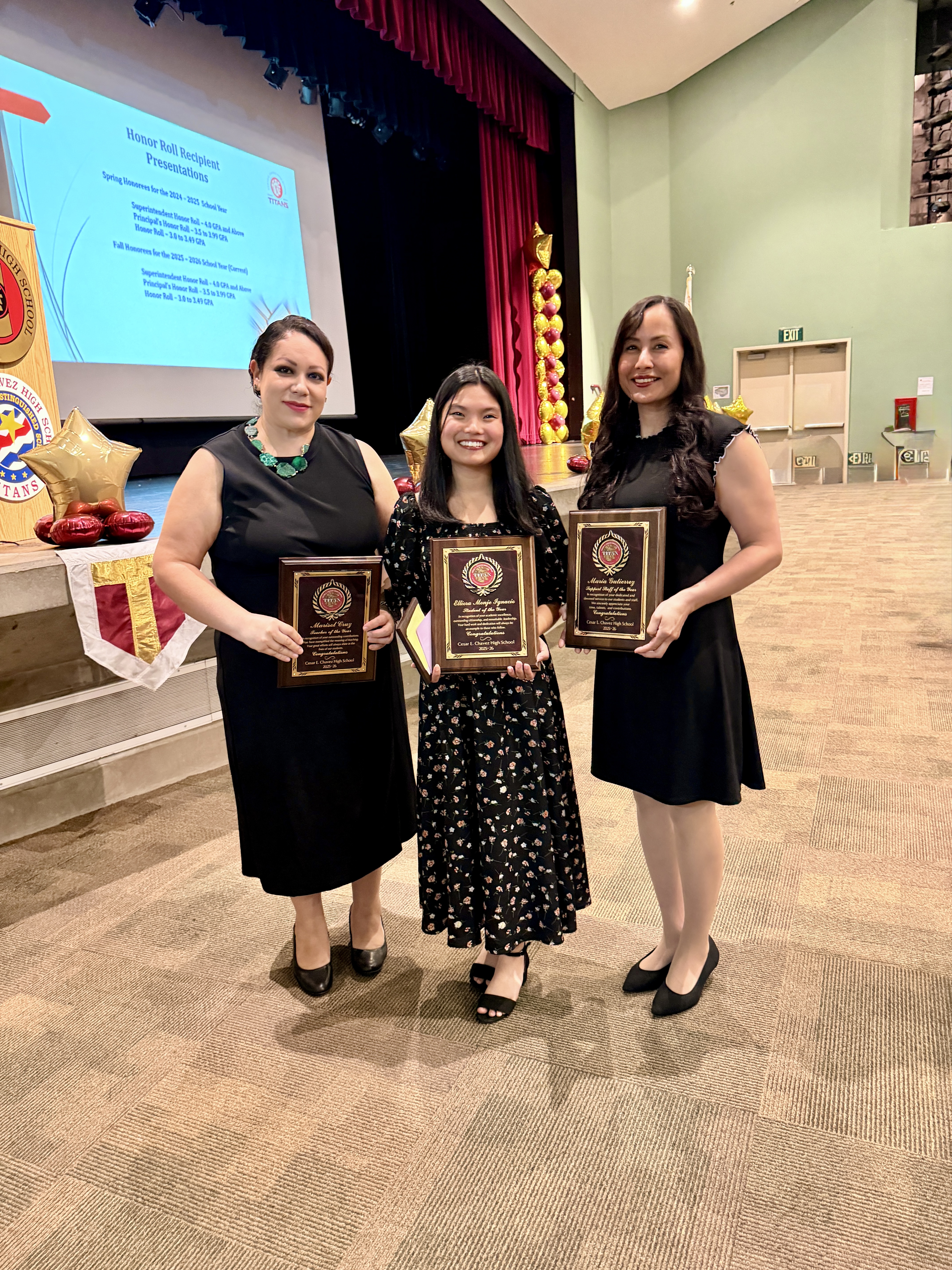 The image shows three women standing together, holding awards or certificates, in what appears to be a stage or auditorium setting with a red curtain backdrop.