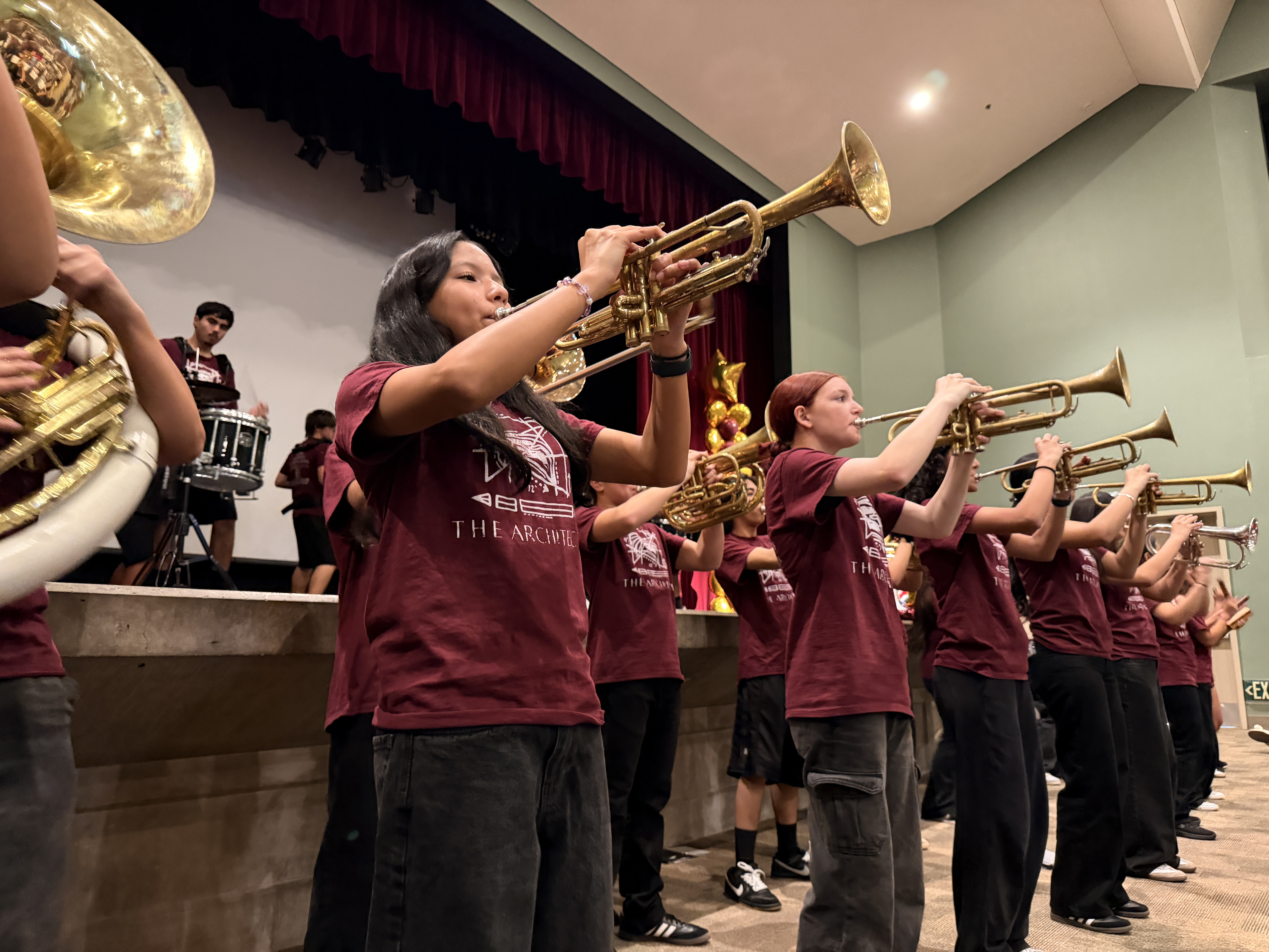 A group of young musicians in maroon uniforms playing brass instruments on a stage with a red curtain backdrop.