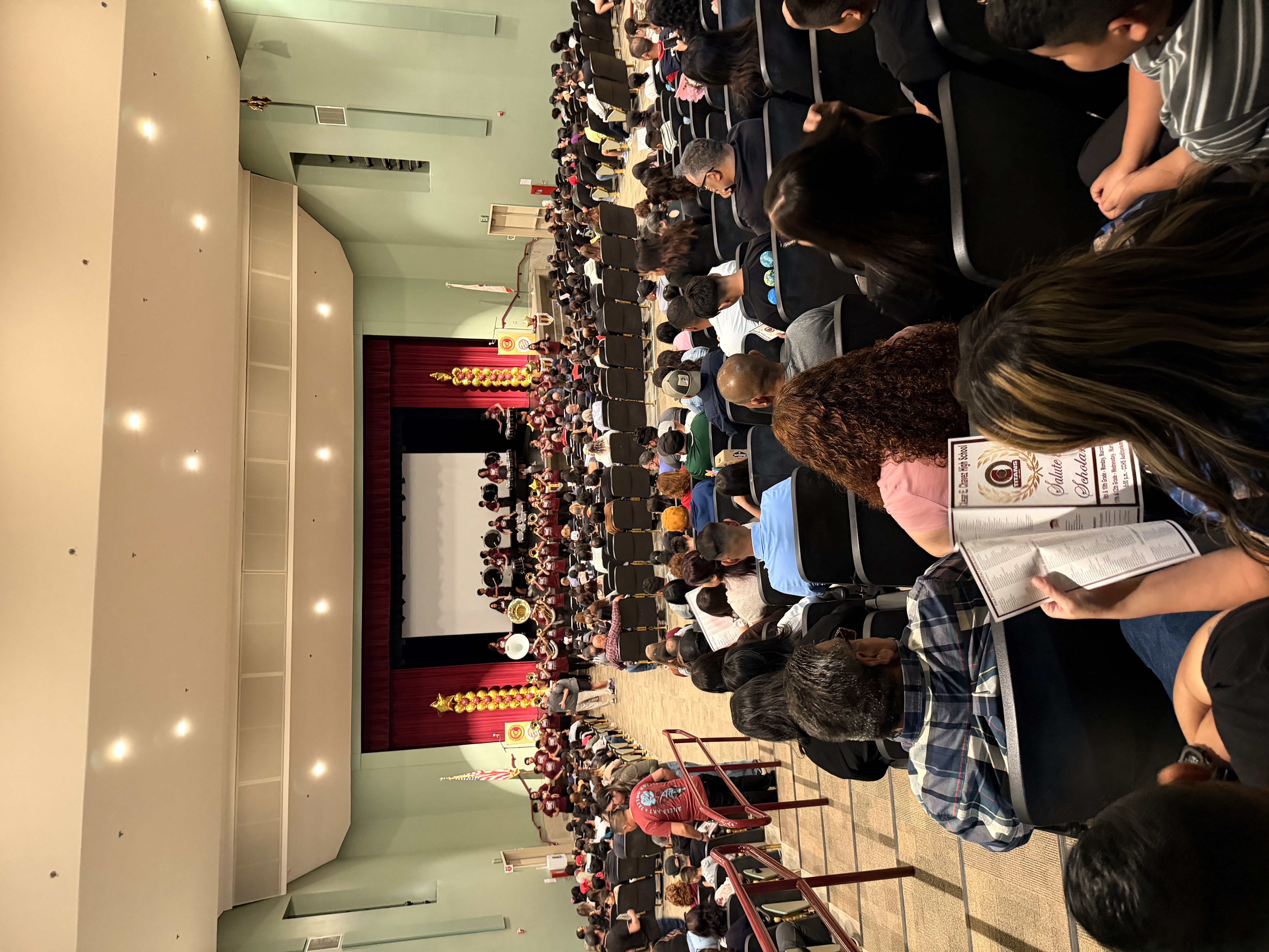 A large auditorium filled with a crowded audience, with a stage and decorations visible in the background.
