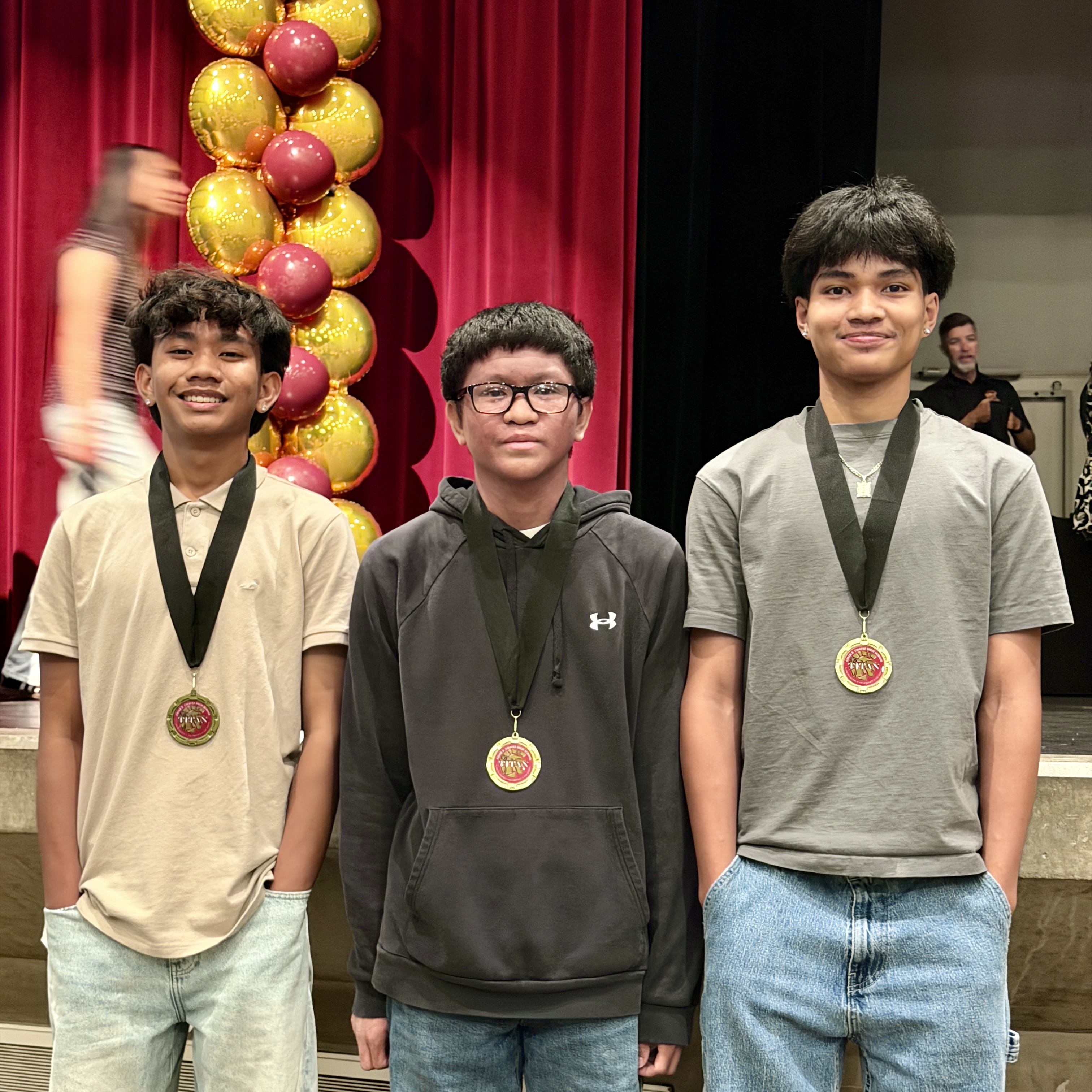 Three young men wearing medals stand in front of a red curtain backdrop with yellow and red balloons.