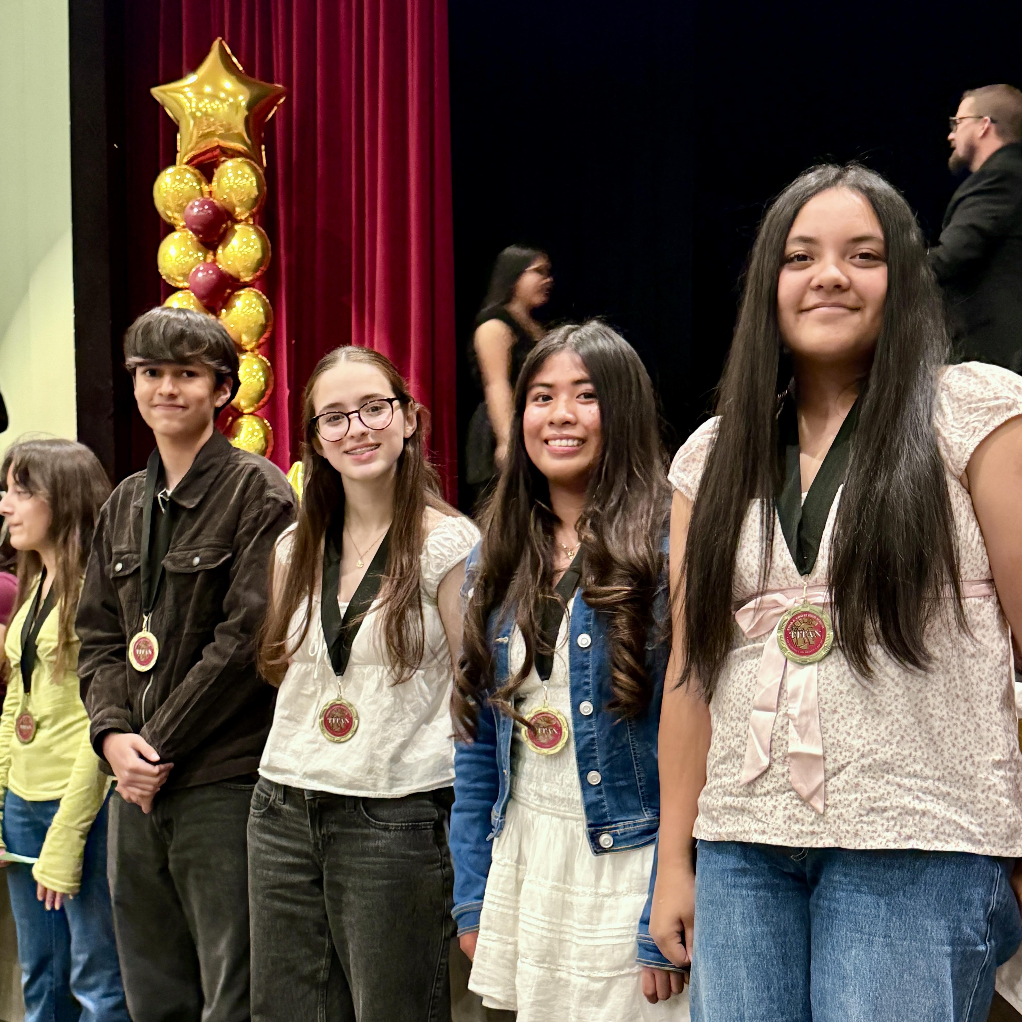 A group of young people, likely students, standing in front of a red curtain backdrop with a star-shaped balloon decoration.