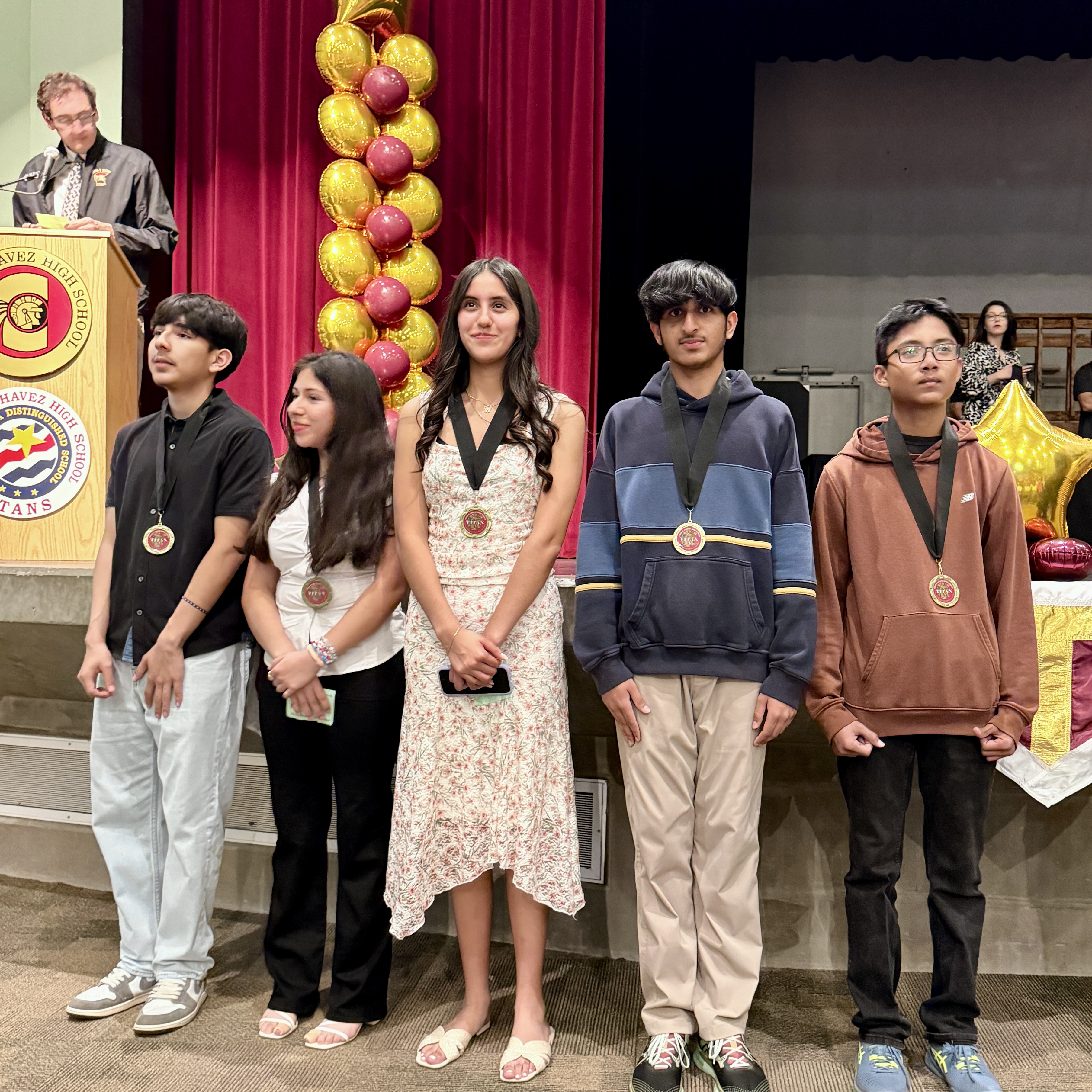 The image shows a group of people standing in front of a red curtain, with a display of yellow apples hanging above them. The people appear to be receiving awards or recognition, as they are holding medals or trophies.