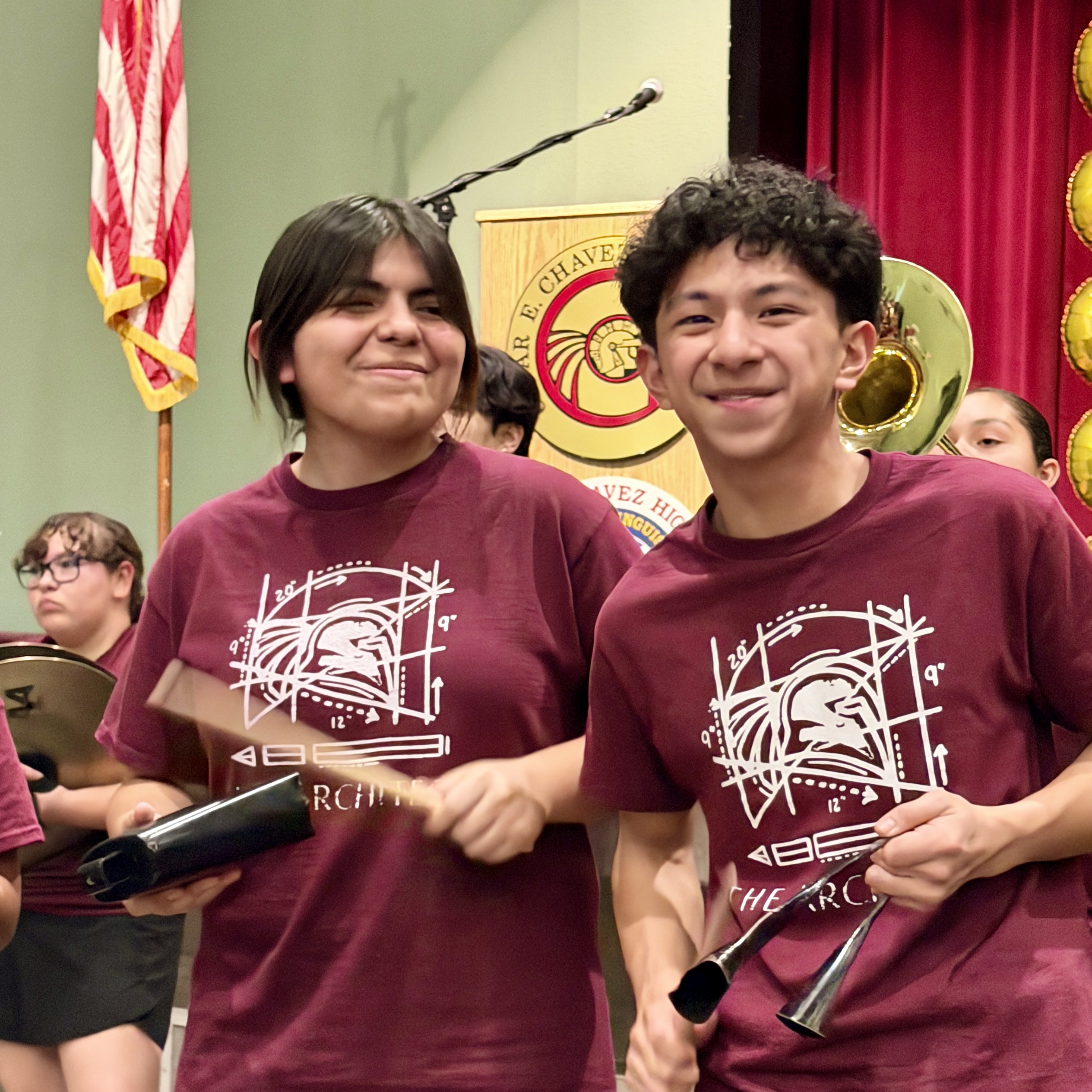 The image shows two young people wearing matching burgundy t-shirts with a graphic design, standing in front of a red curtain backdrop with a flag and emblem visible.