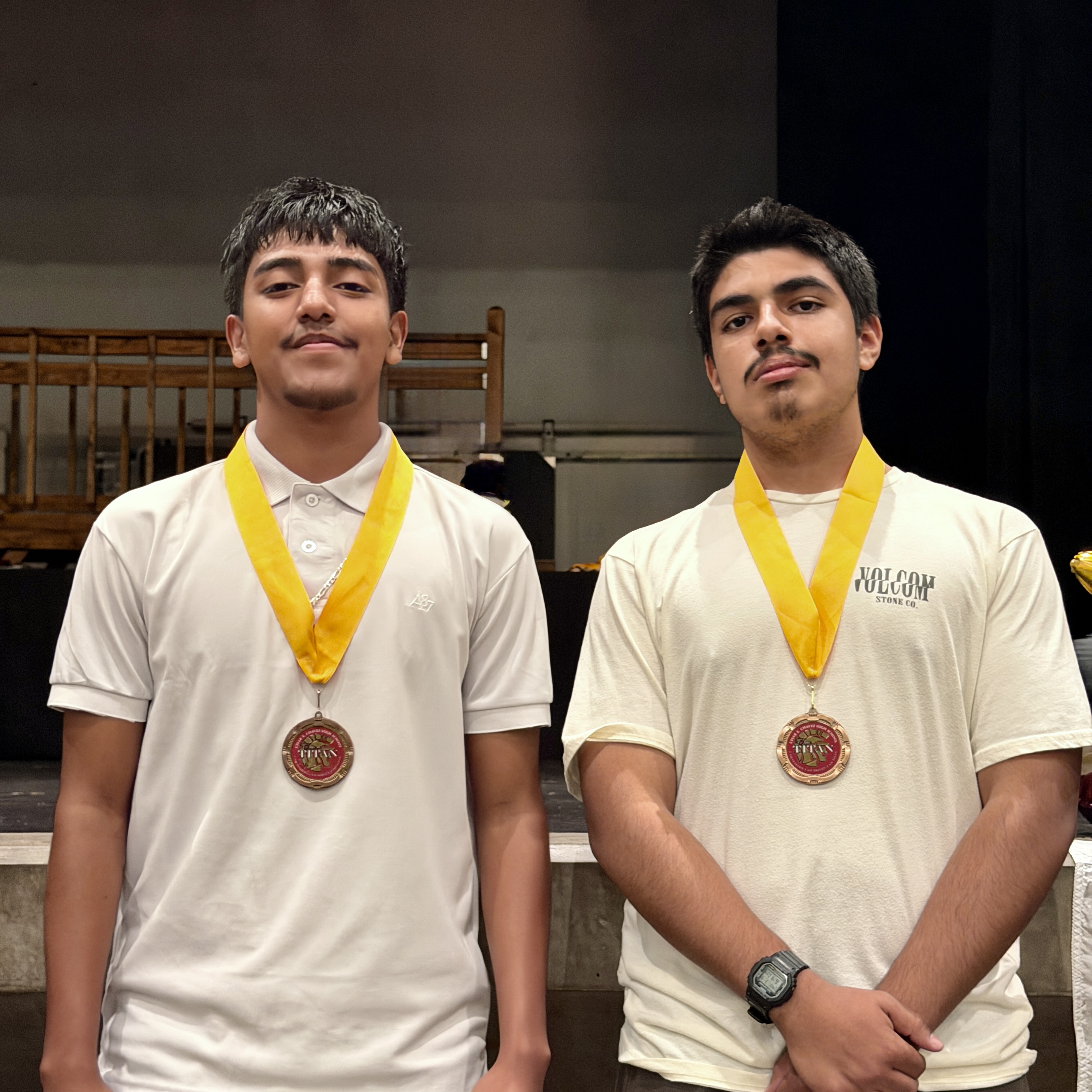 Two young men wearing white shirts and yellow medals around their necks, standing in what appears to be a stage or performance area with a wooden railing in the background.