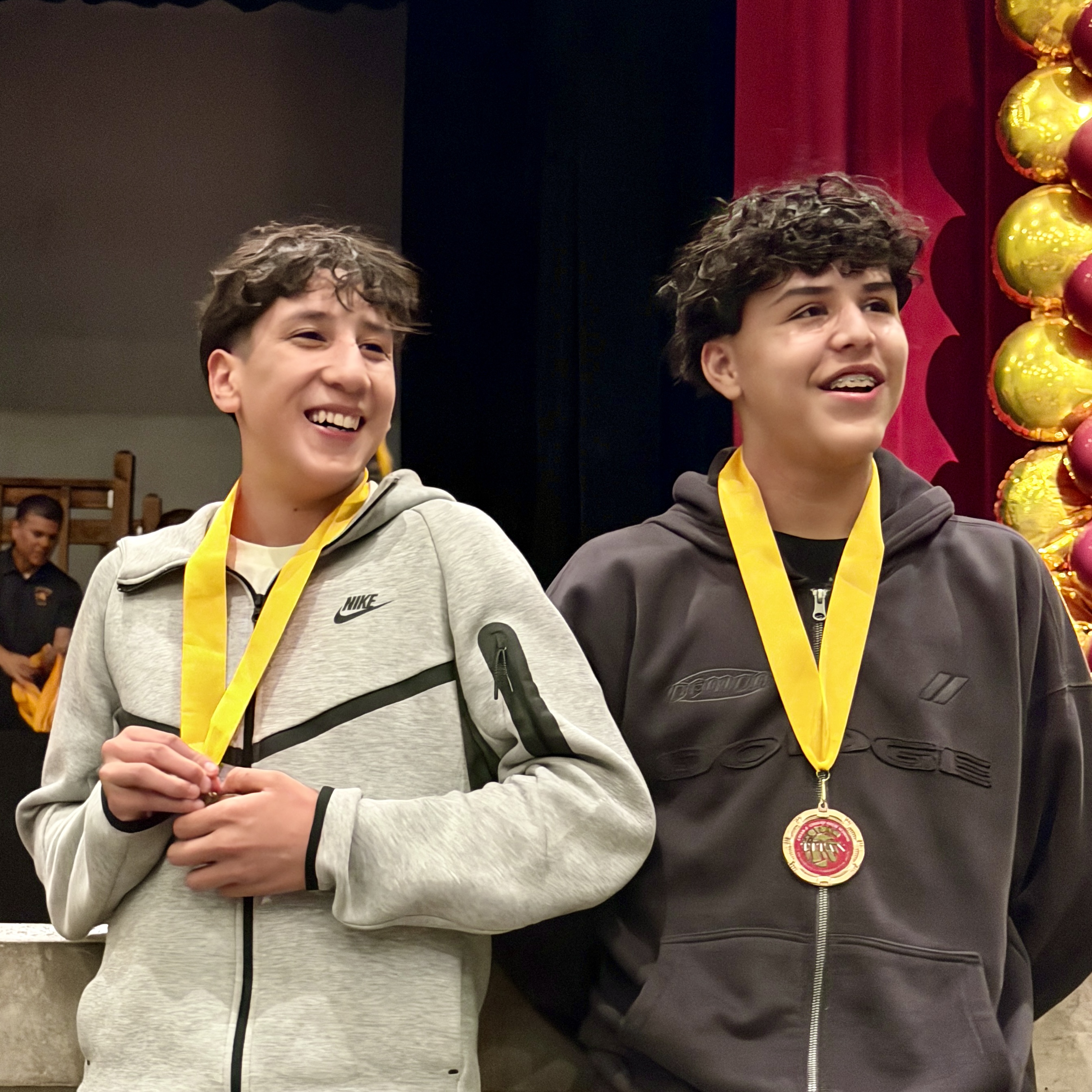 Two young men with curly hair are standing together, wearing gray and black hoodies, and holding gold medals around their necks. They appear to be in a stage or performance setting, with a red curtain visible in the background.