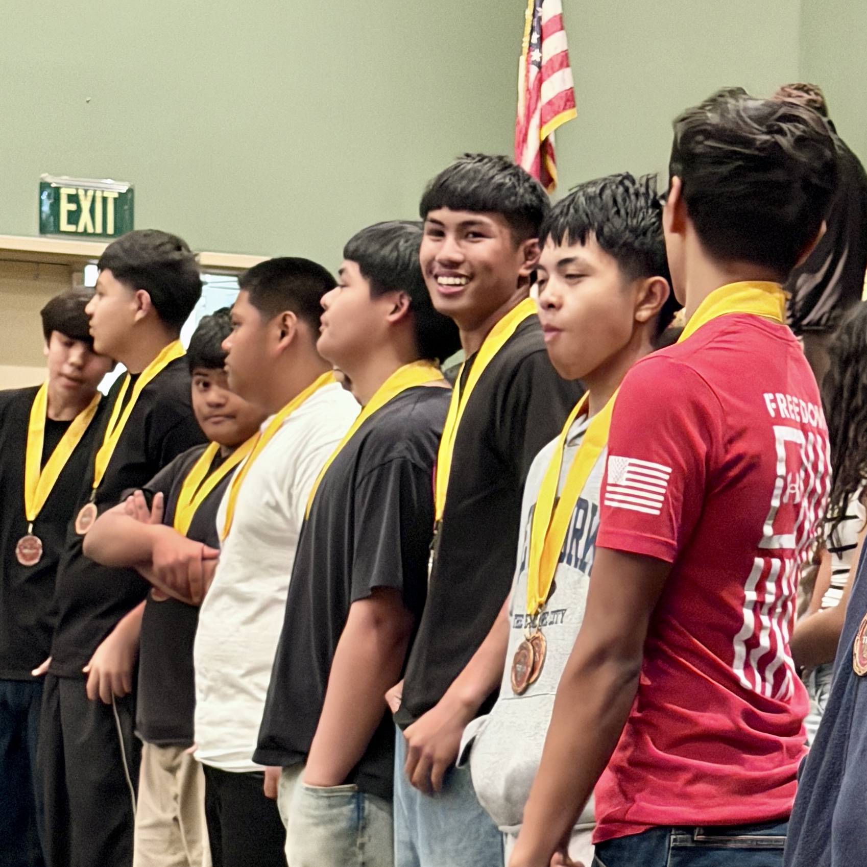 A group of young boys wearing yellow and black shirts, standing in a row and smiling, with an American flag and an exit sign visible in the background.