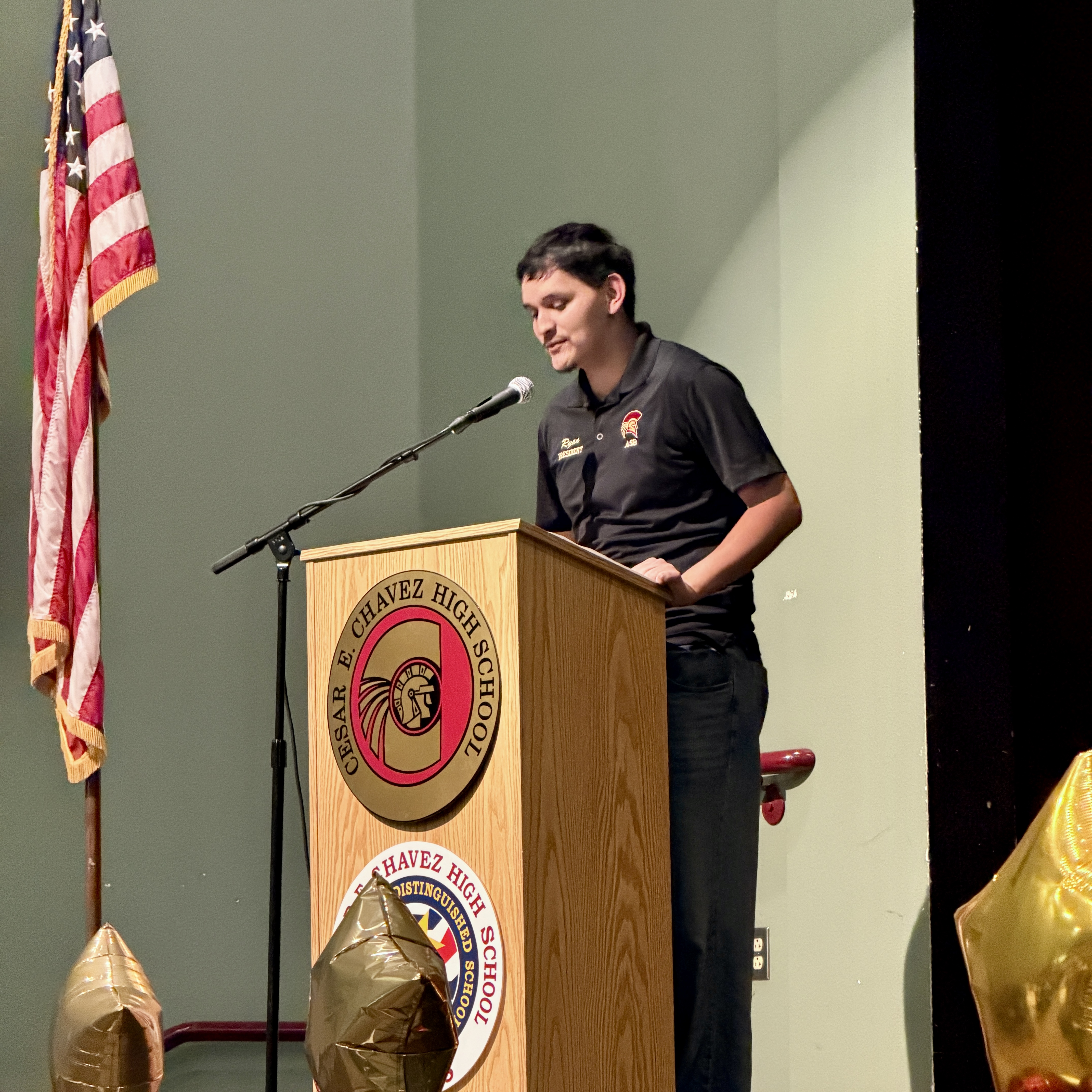 A person in a police uniform is standing at a podium with a microphone, in front of an American flag and a logo on the wall behind them.