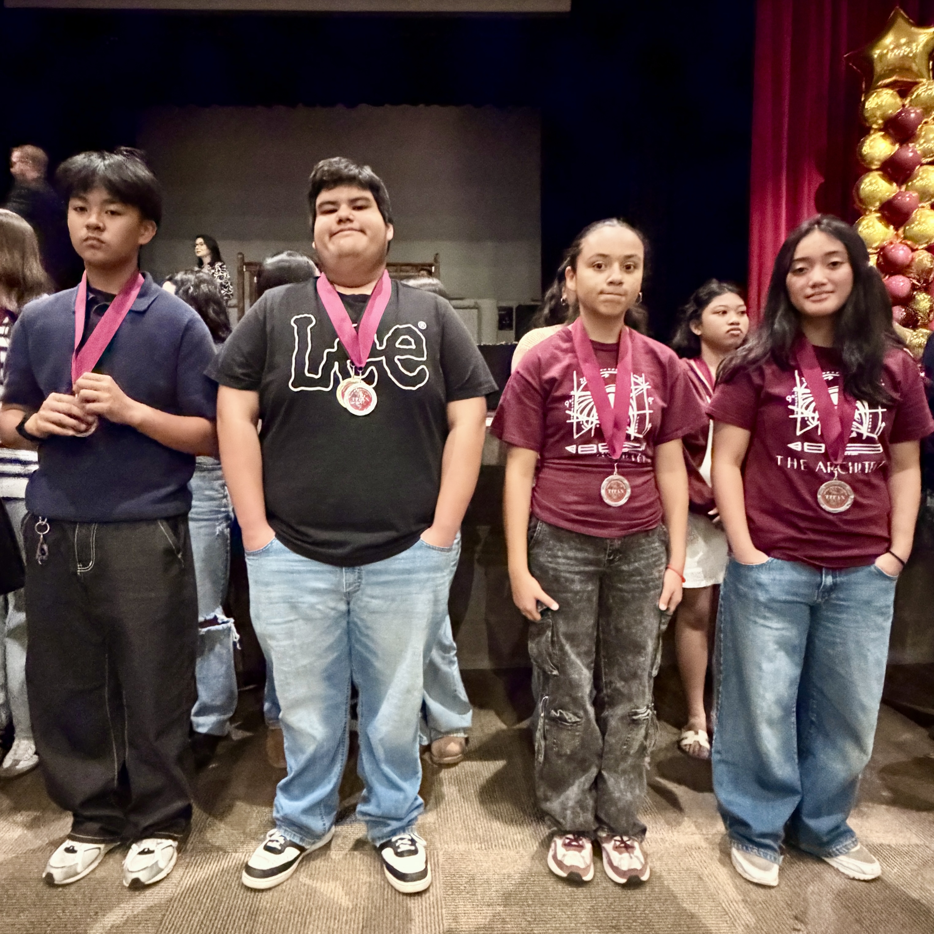The image shows a group of young people, likely students, standing together in what appears to be a school or community setting, with decorations and banners visible in the background.