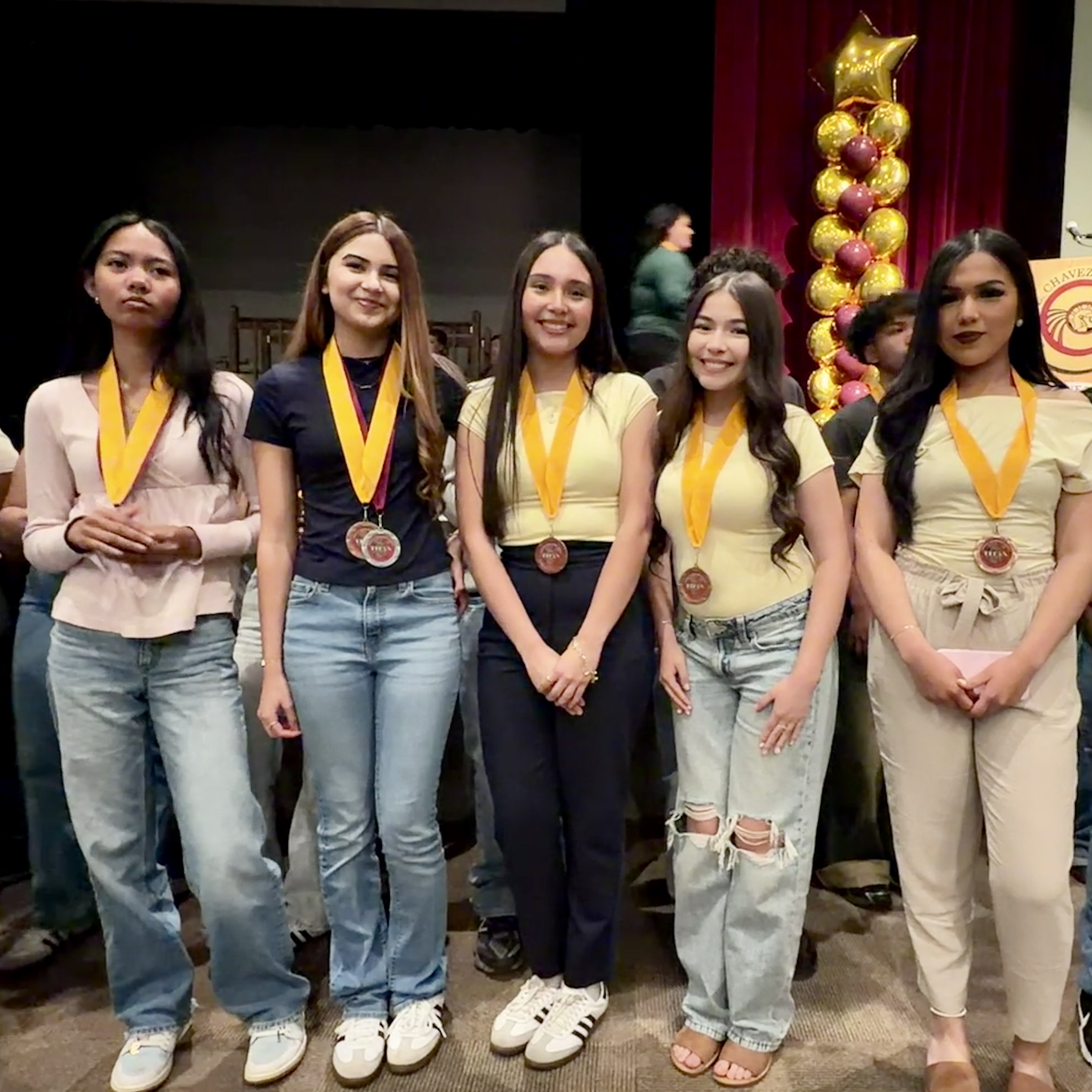 A group of young women in casual attire, wearing yellow medals, stand in front of a red curtain backdrop adorned with golden star decorations.