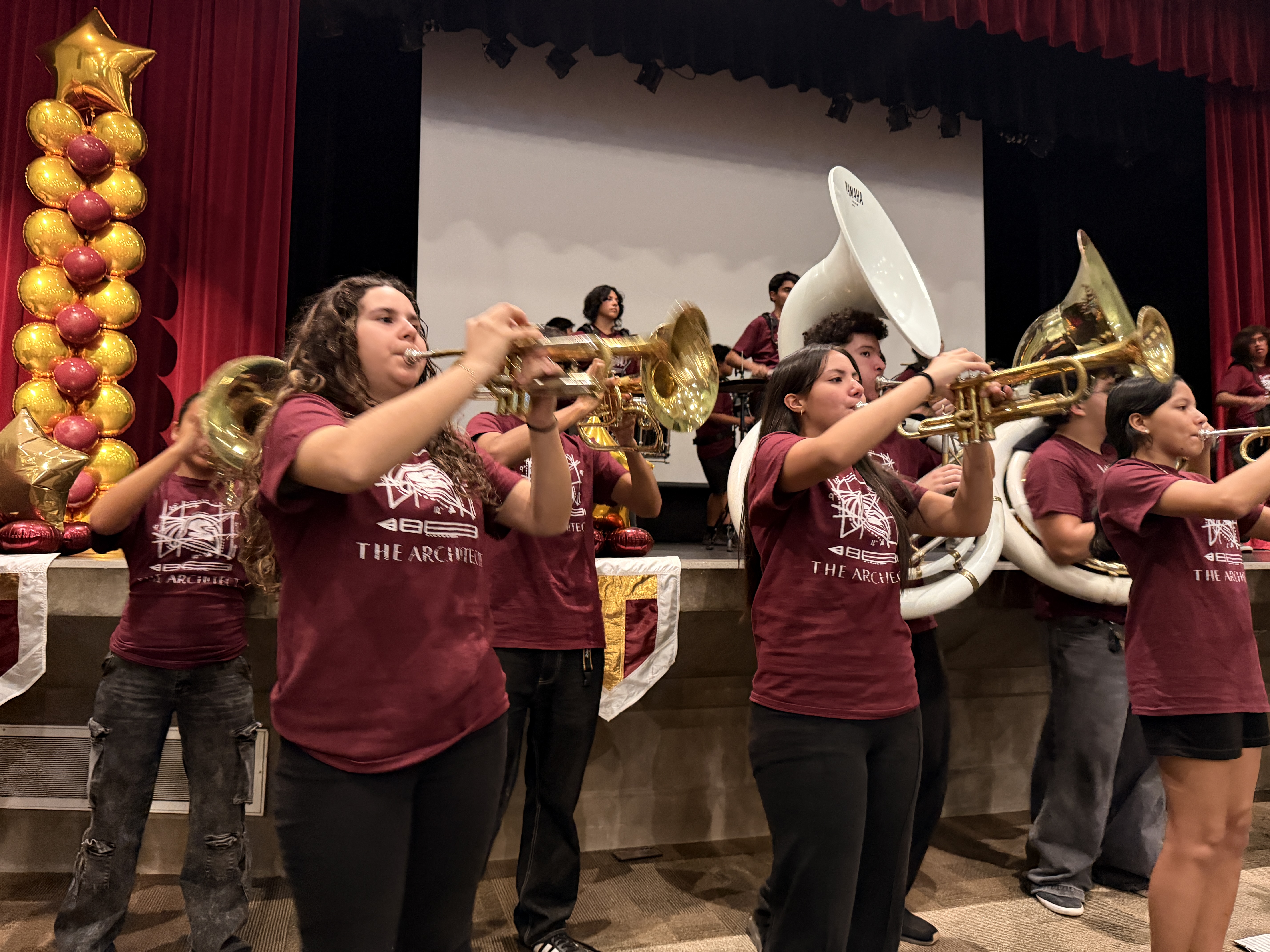 A group of people wearing maroon shirts are playing musical instruments on a stage with a red curtain backdrop and various decorations.