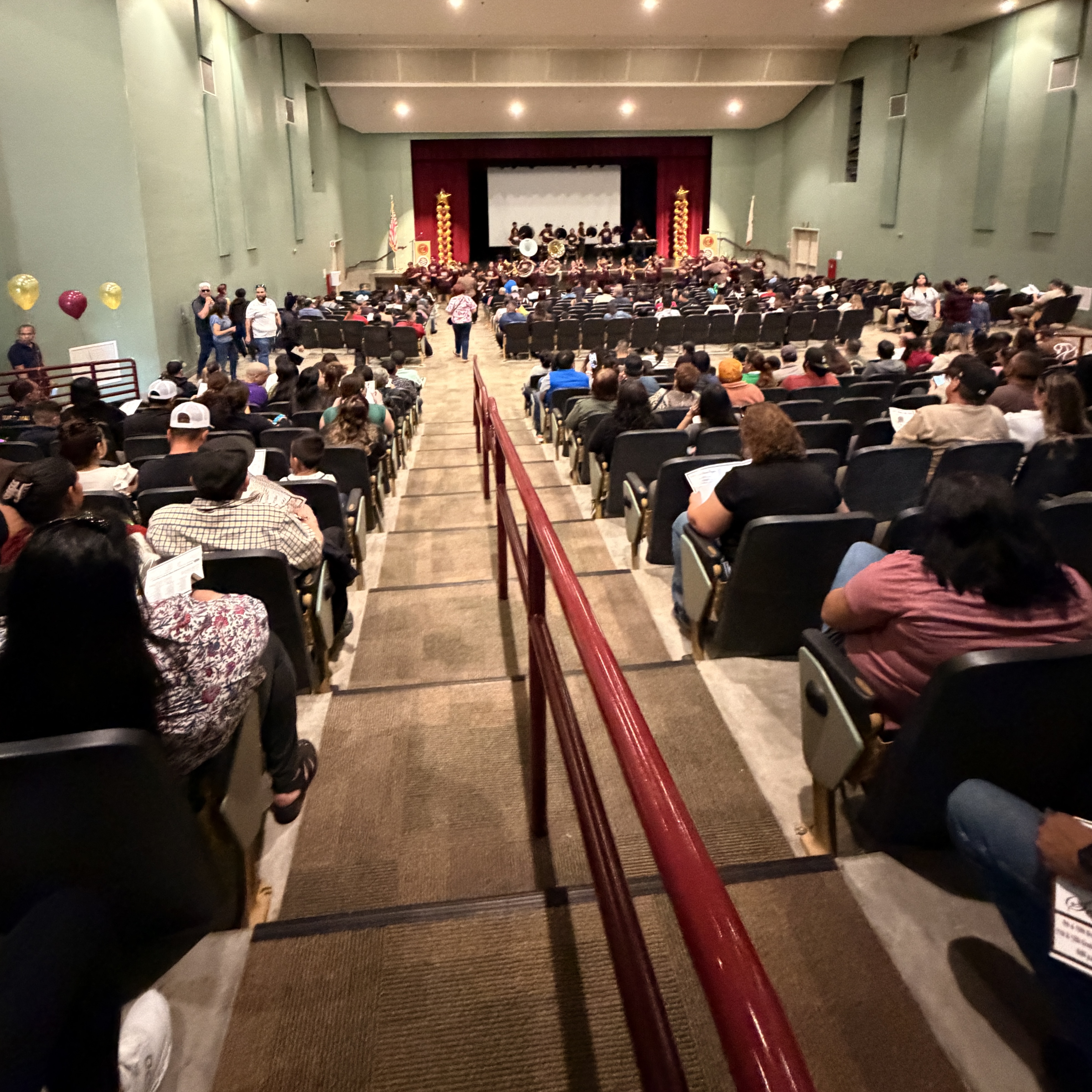 A large auditorium filled with rows of people seated, facing a stage with decorations and lighting, suggesting a performance or event taking place.