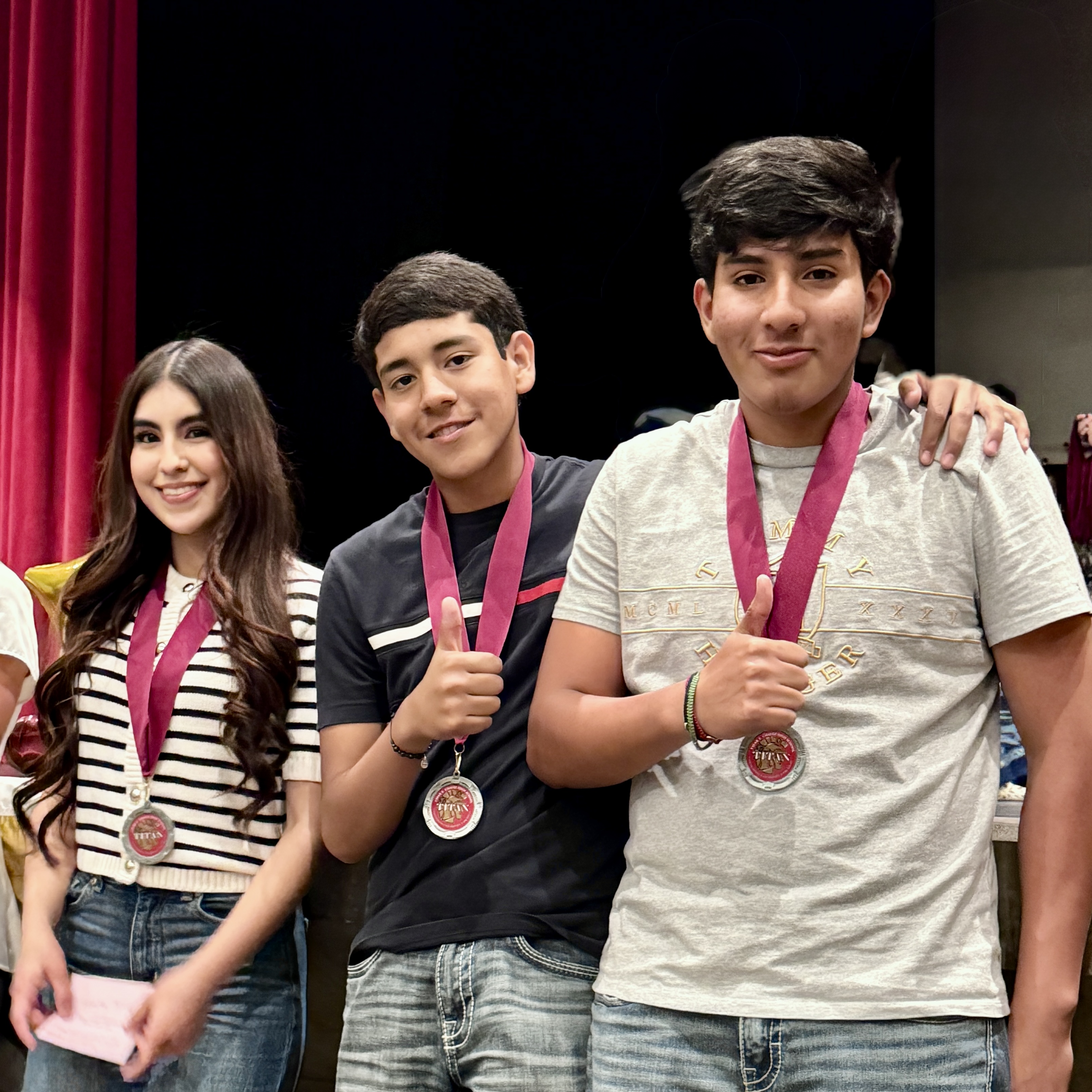 Three young people, two males and one female, are standing together and holding medals around their necks. They appear to be in a performance or competition setting, with a red curtain visible in the background.