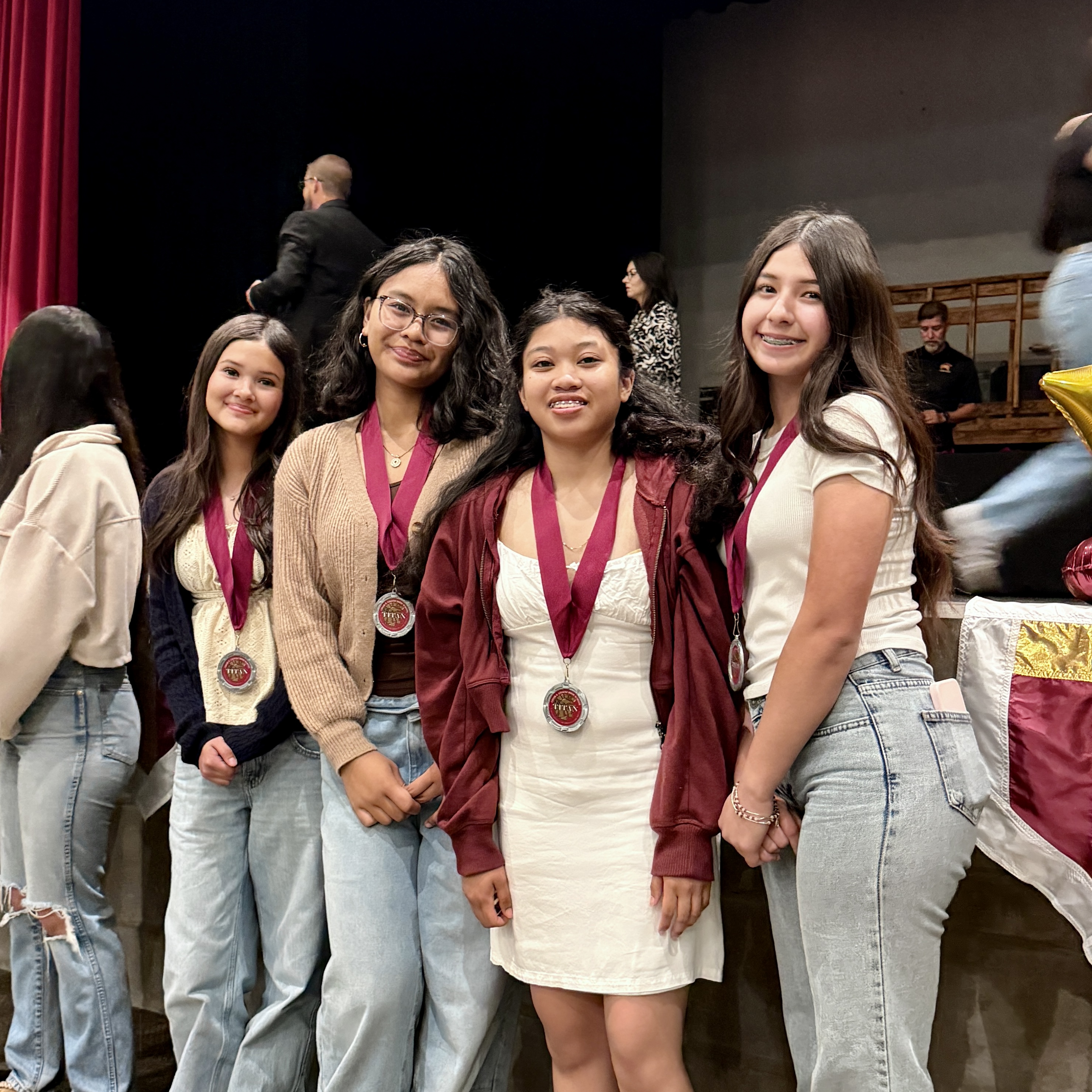 A group of young women, some wearing medals, stand together in a theater setting with a red curtain in the background.