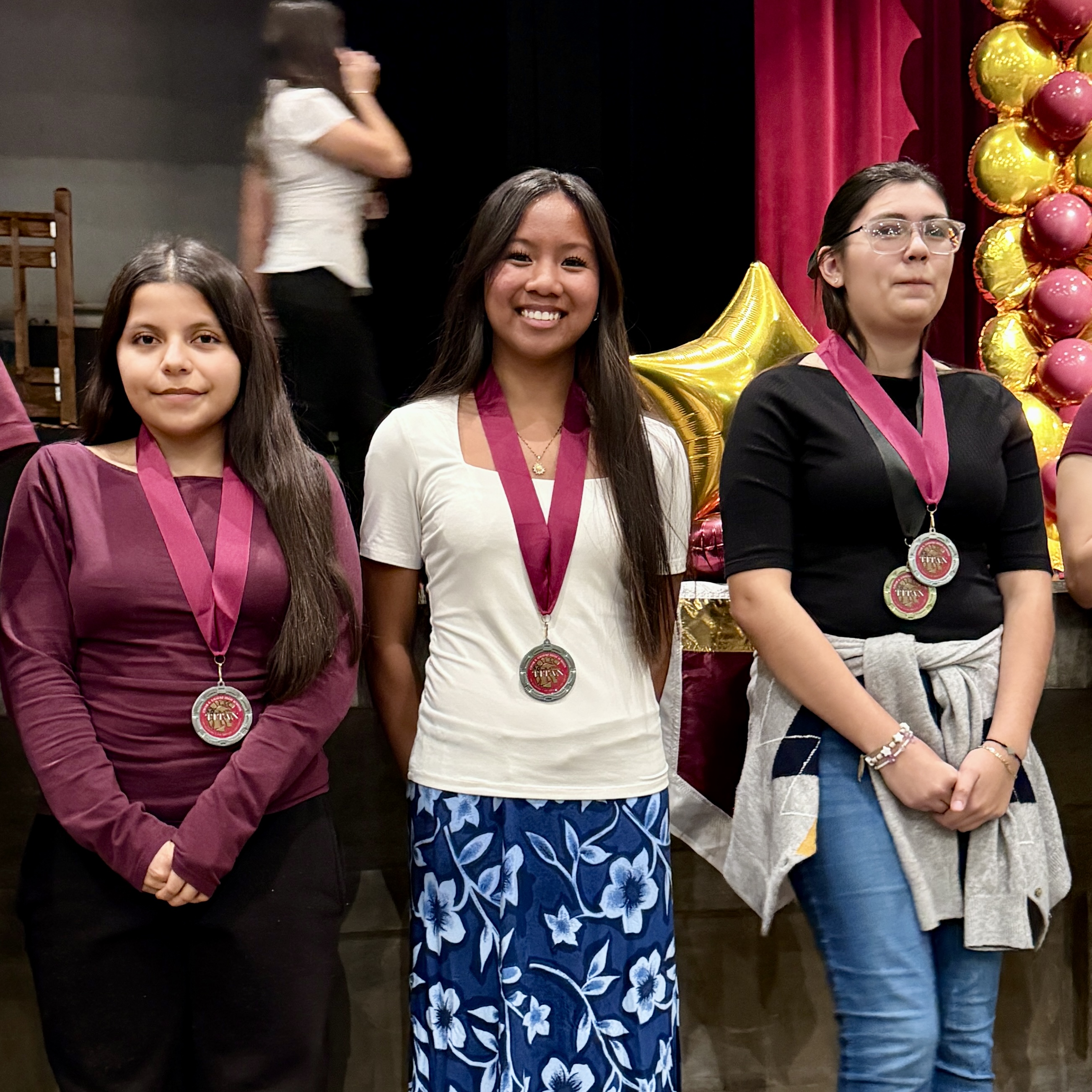 Three young women wearing medals and colorful attire stand in front of a red curtain backdrop, with balloons visible in the background.