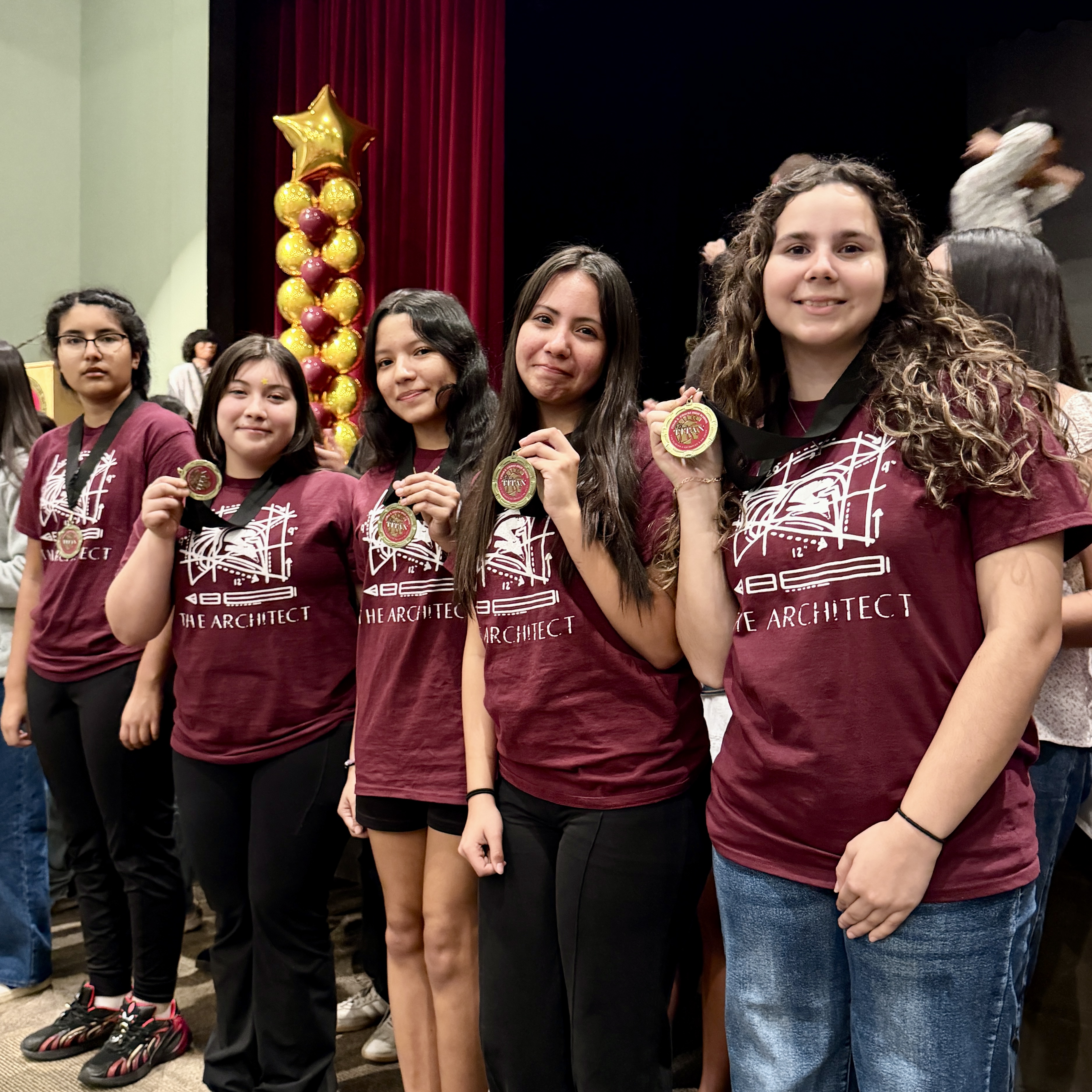 A group of young women wearing matching burgundy t-shirts with the text "ARCHITECT" stand together in front of a red curtain, holding various awards and decorations.