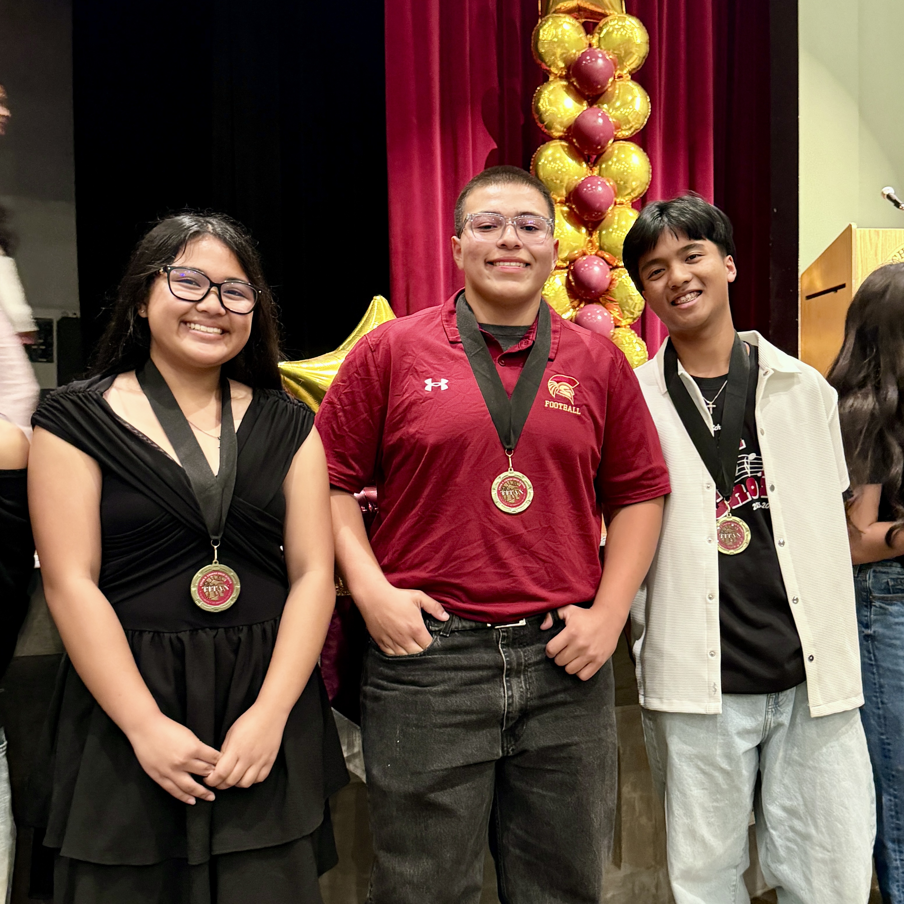 The image shows three young people standing in front of a red curtain with yellow balloons behind them. The person in the middle is wearing a red shirt and holding a medal, while the other two individuals are wearing casual clothing.