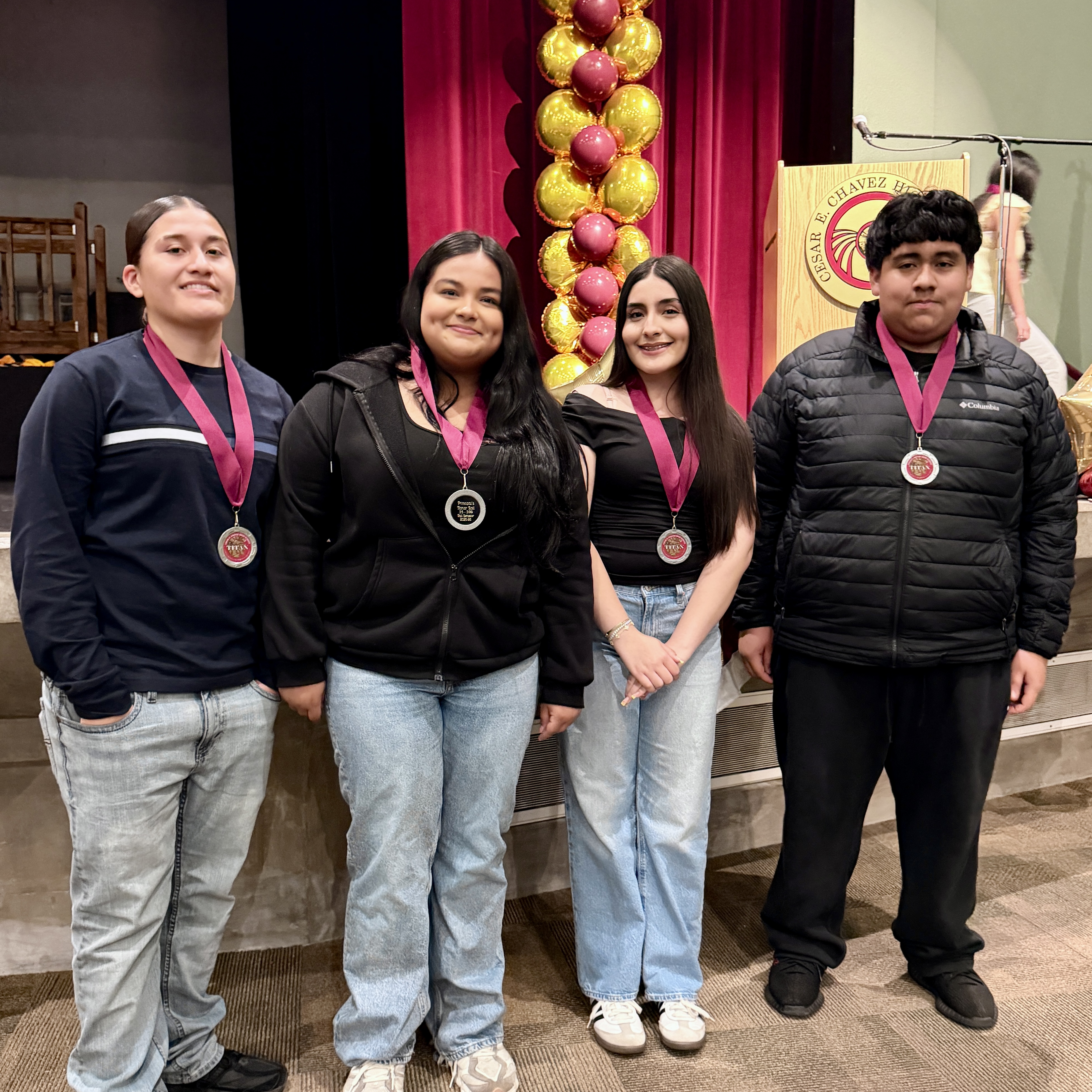 Four individuals, two women and two men, are standing together in a room with a red curtain backdrop and decorative balloons. They are all wearing medals around their necks, suggesting they have recently participated in some kind of event or competition.