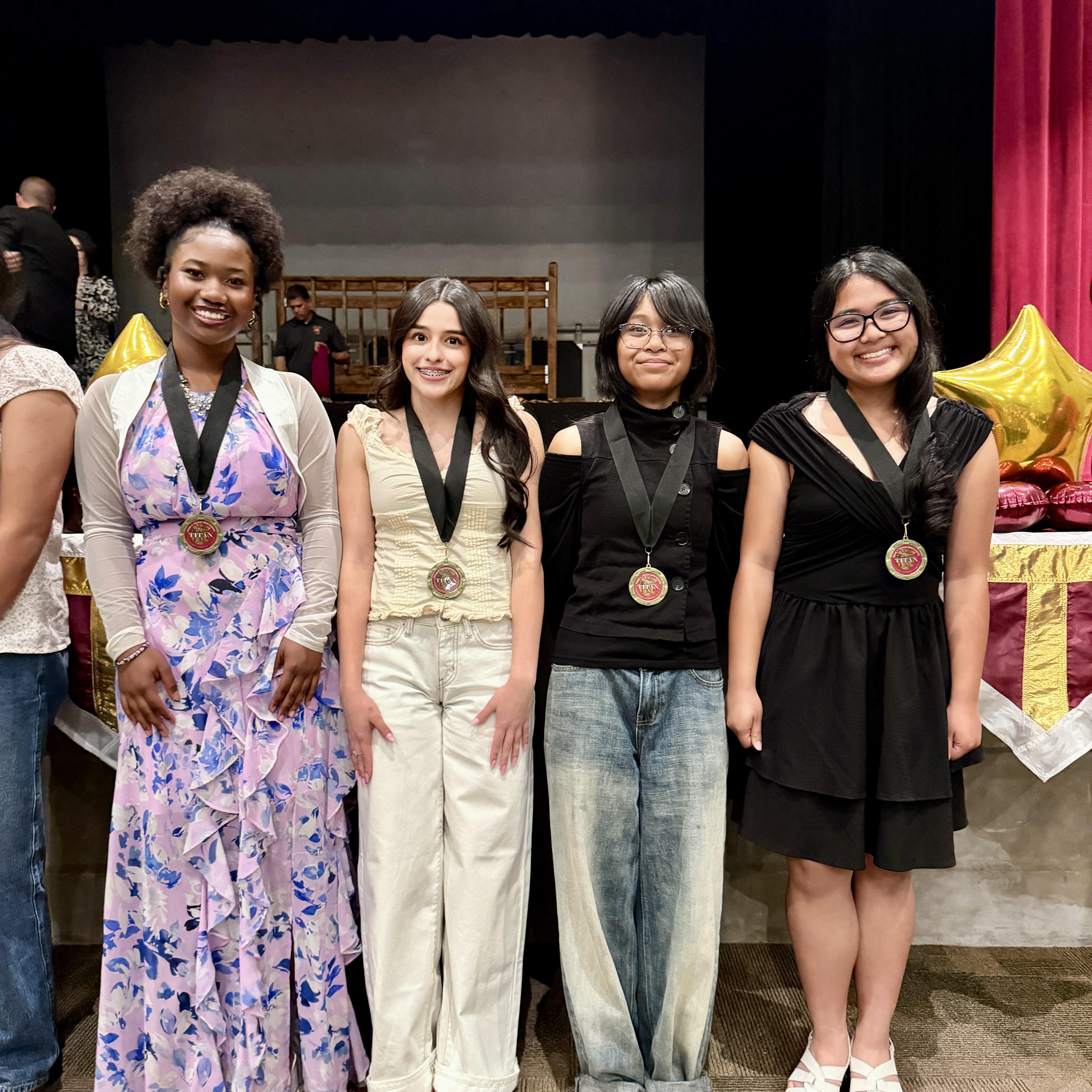 The image shows a group of four young women standing together on a stage, dressed in various stylish outfits and holding medals or awards.