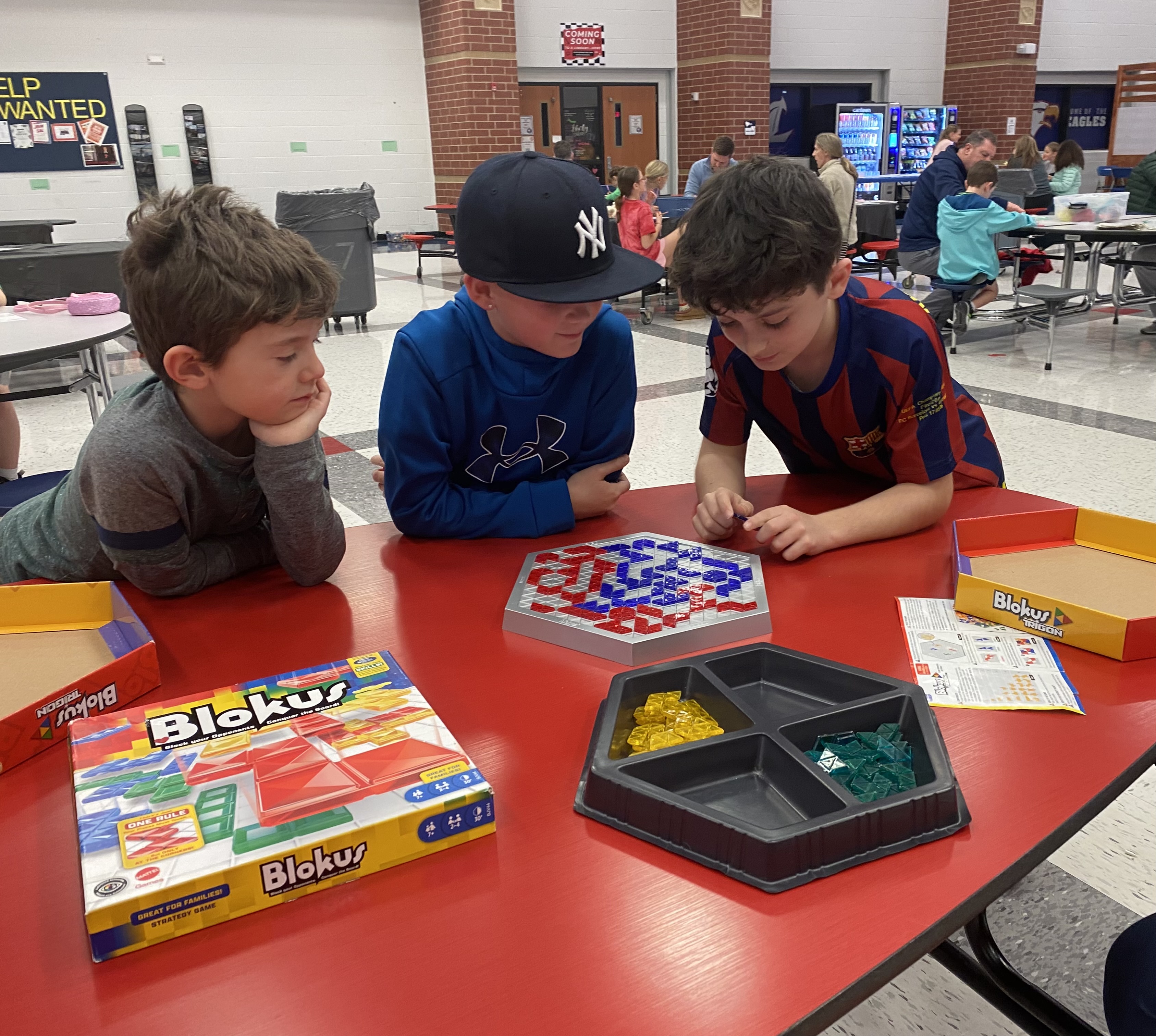 Three young boys are playing a board game together at a table in what appears to be a school cafeteria or common area, with various other students and activities visible in the background.