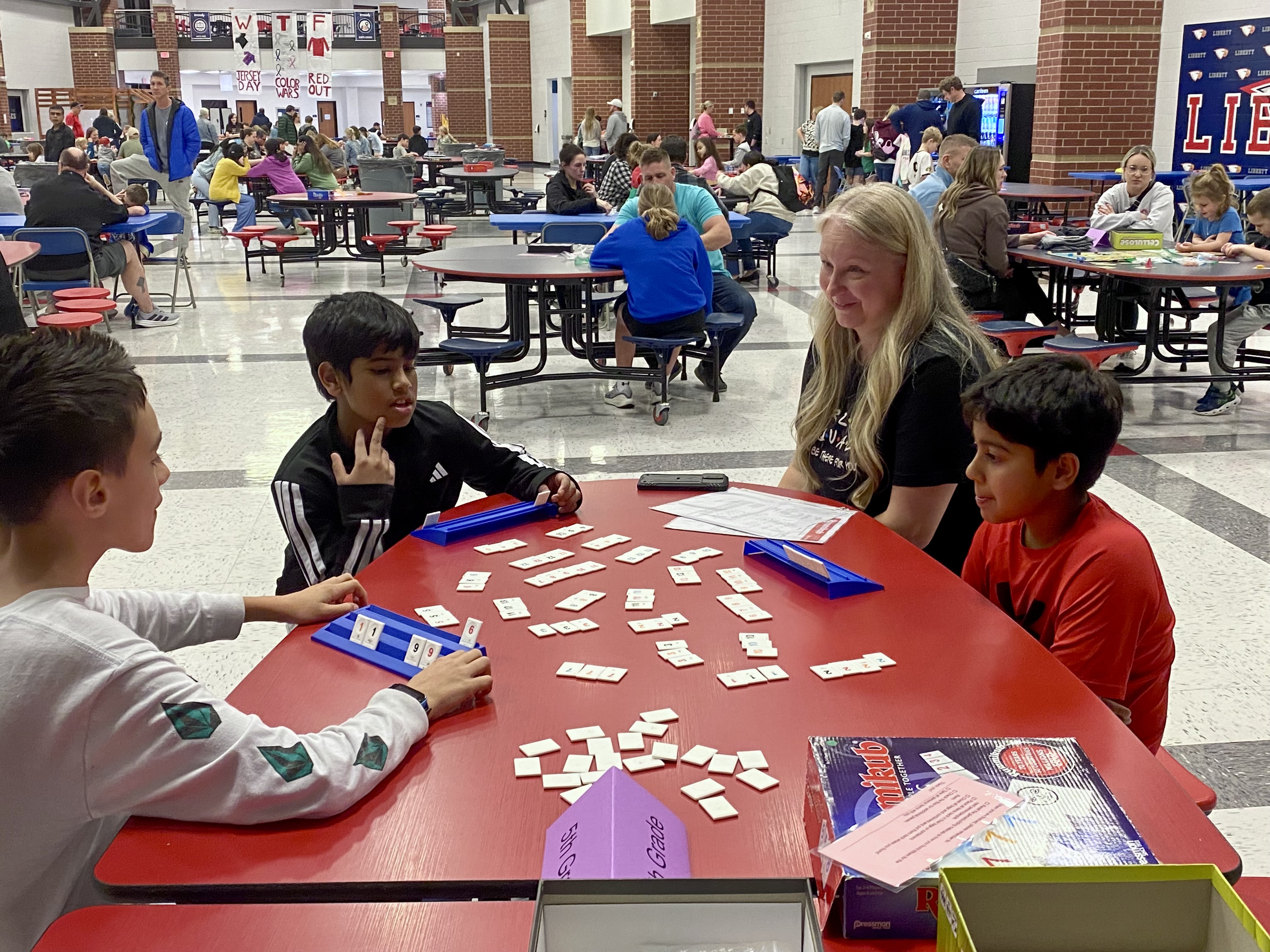 A group of students are gathered around a table, engaged in what appears to be a collaborative activity or game, while the background shows a busy school cafeteria setting with other students and staff present.