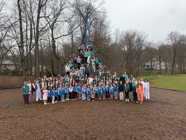 A large group of children in colorful outfits poses on a playground with a climbing structure in the background and wood chips in the foreground.