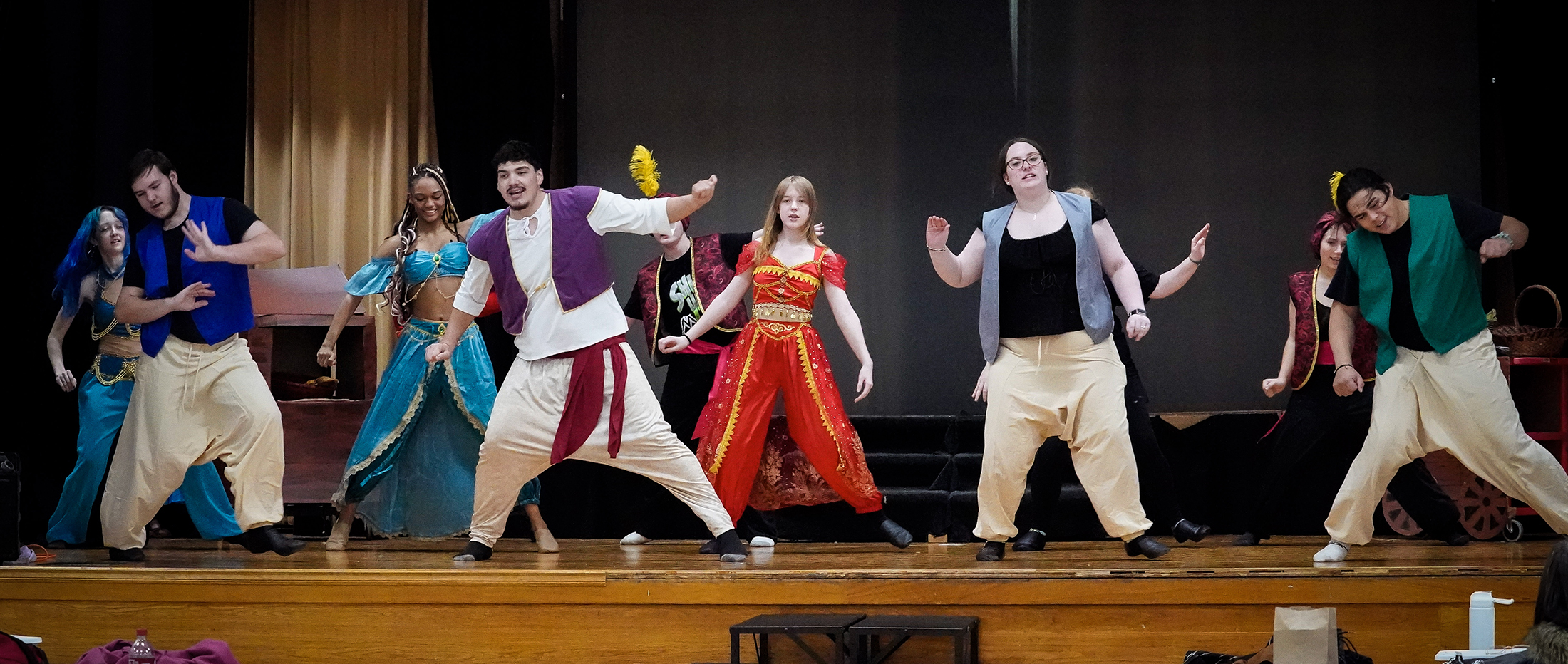 A group of performers in colorful, vibrant costumes are dancing on a stage with a dark background.