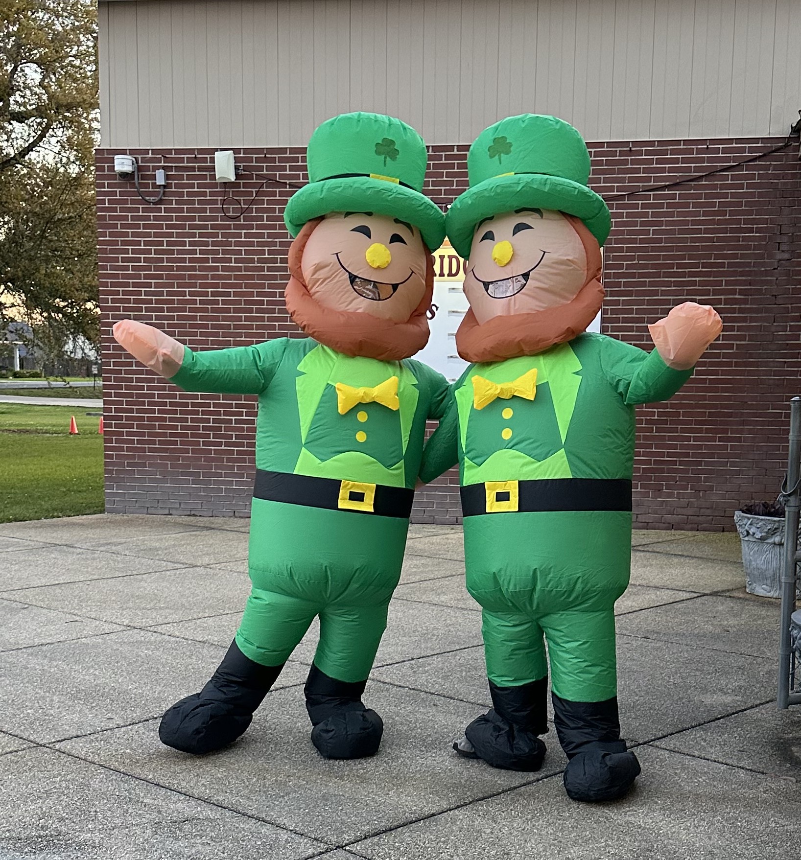 Two inflatable leprechauns stand on a paved walkway in front of a brick building.