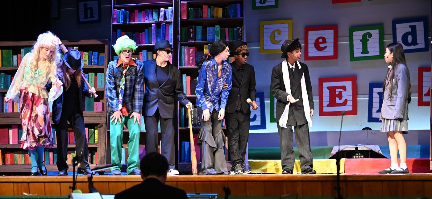The image shows a group of people, likely performers, standing on a stage in front of a colorful backdrop with bookshelves and various decorative elements.