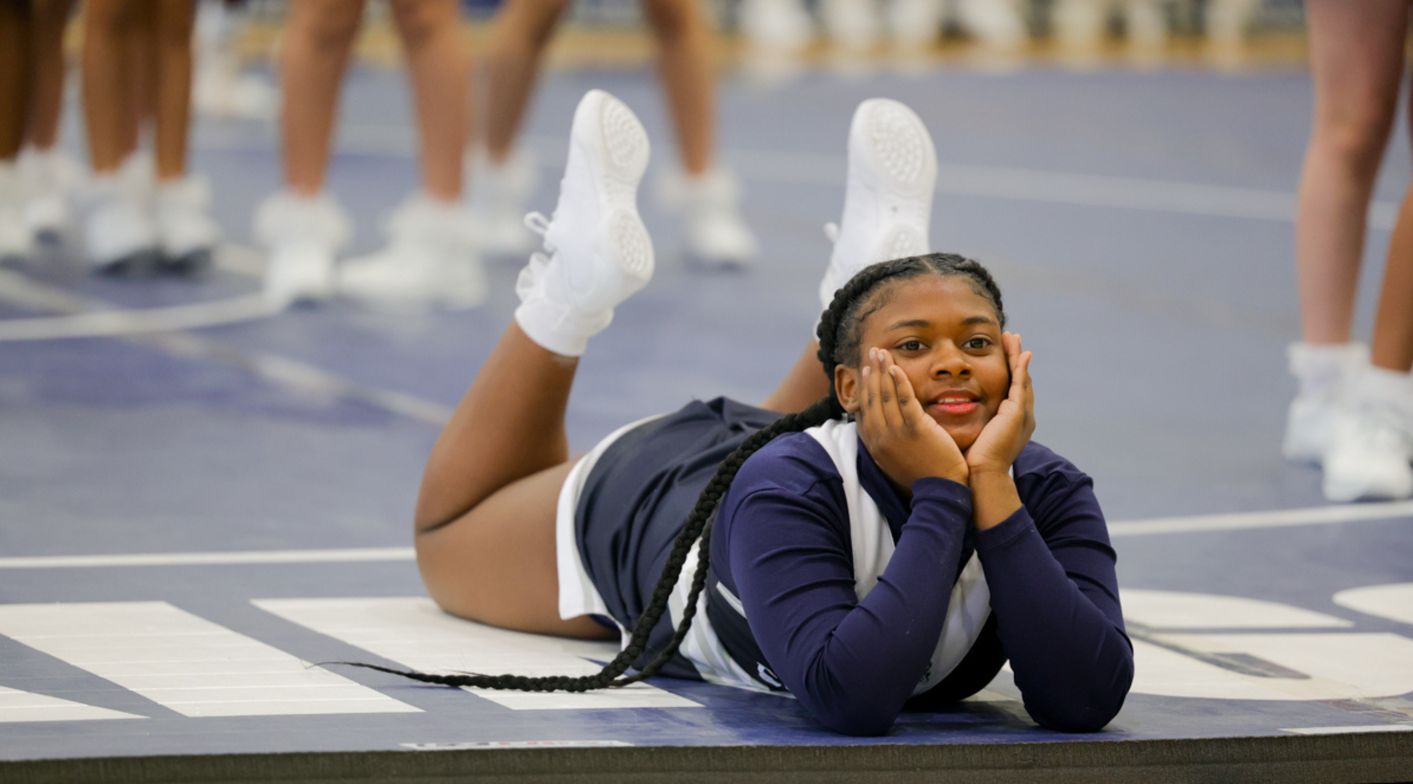 A young woman in a navy blue outfit sits on a white floor, with a backdrop of wooden dance studio walls and floors.