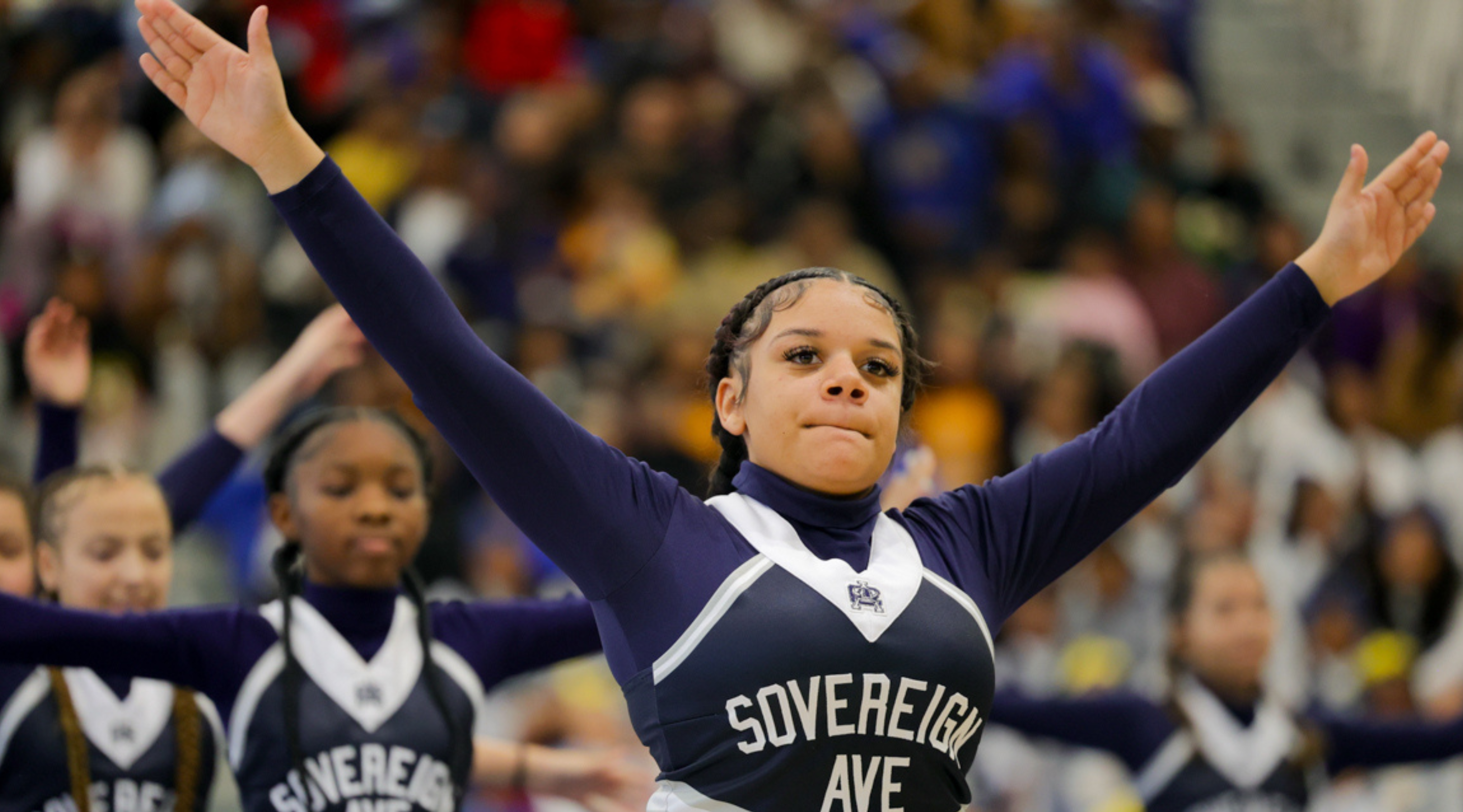 A young woman in a navy blue uniform with "Sovereign Ave" printed on it stands with her arms raised, surrounded by a crowd of people in the background.