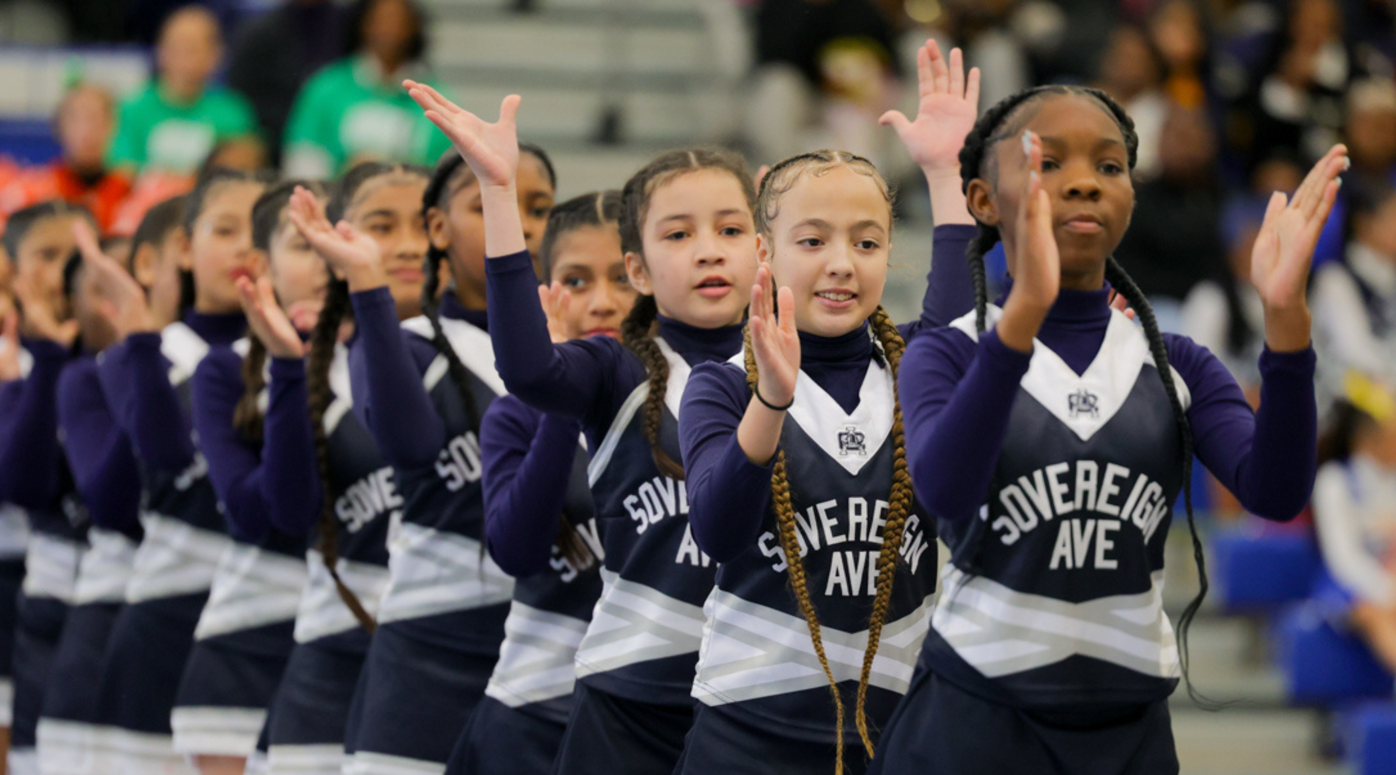 A group of young students wearing matching uniforms, with some raising their hands, appears to be participating in a performance or event in a large indoor venue.