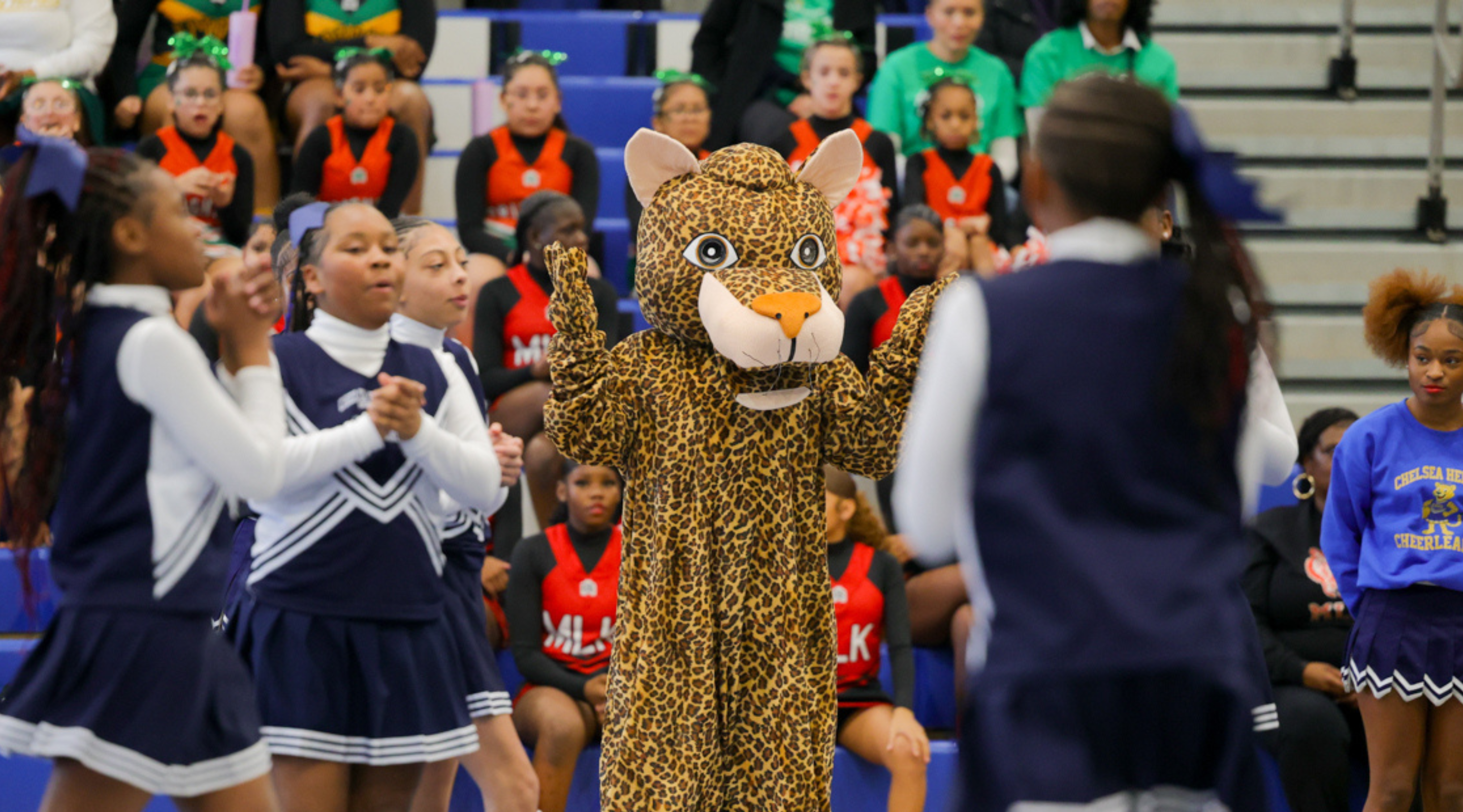 A group of cheerleaders in uniform are gathered around a person dressed in a leopard-print costume, likely a mascot or character, in what appears to be a gymnasium or sports arena setting.