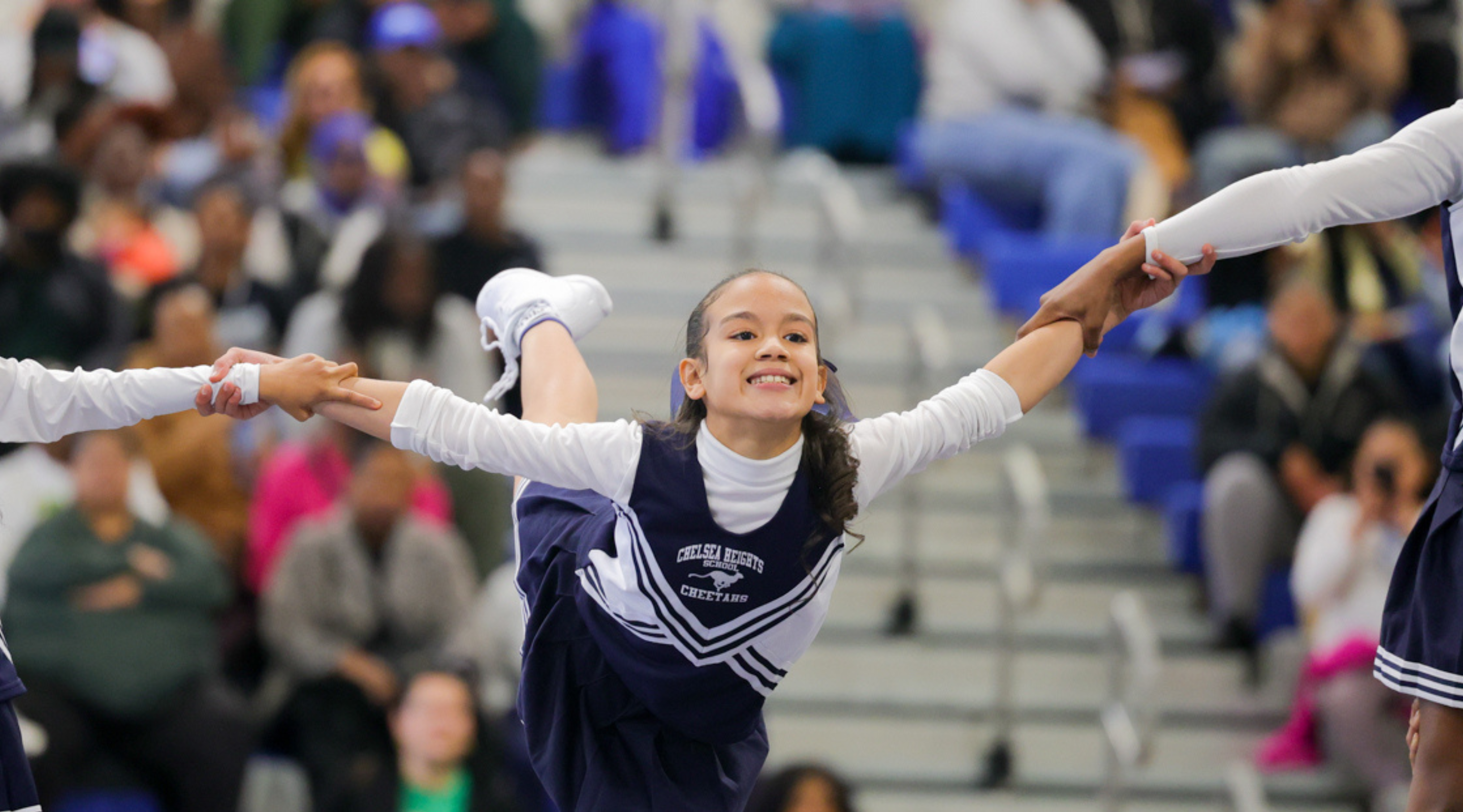 A young cheerleader in a navy and white uniform is enthusiastically raising her arms, surrounded by a crowd of spectators in the background.