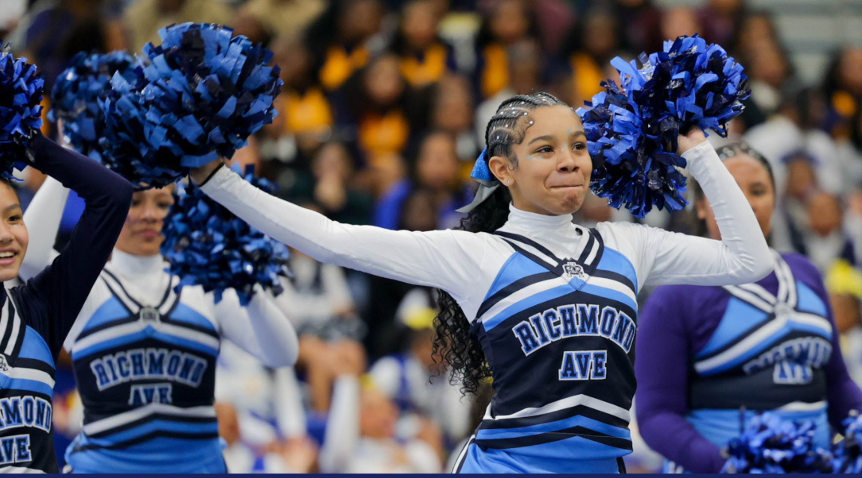 A group of cheerleaders in blue and white uniforms, holding blue and white pom-poms, are performing on a sports field or arena.