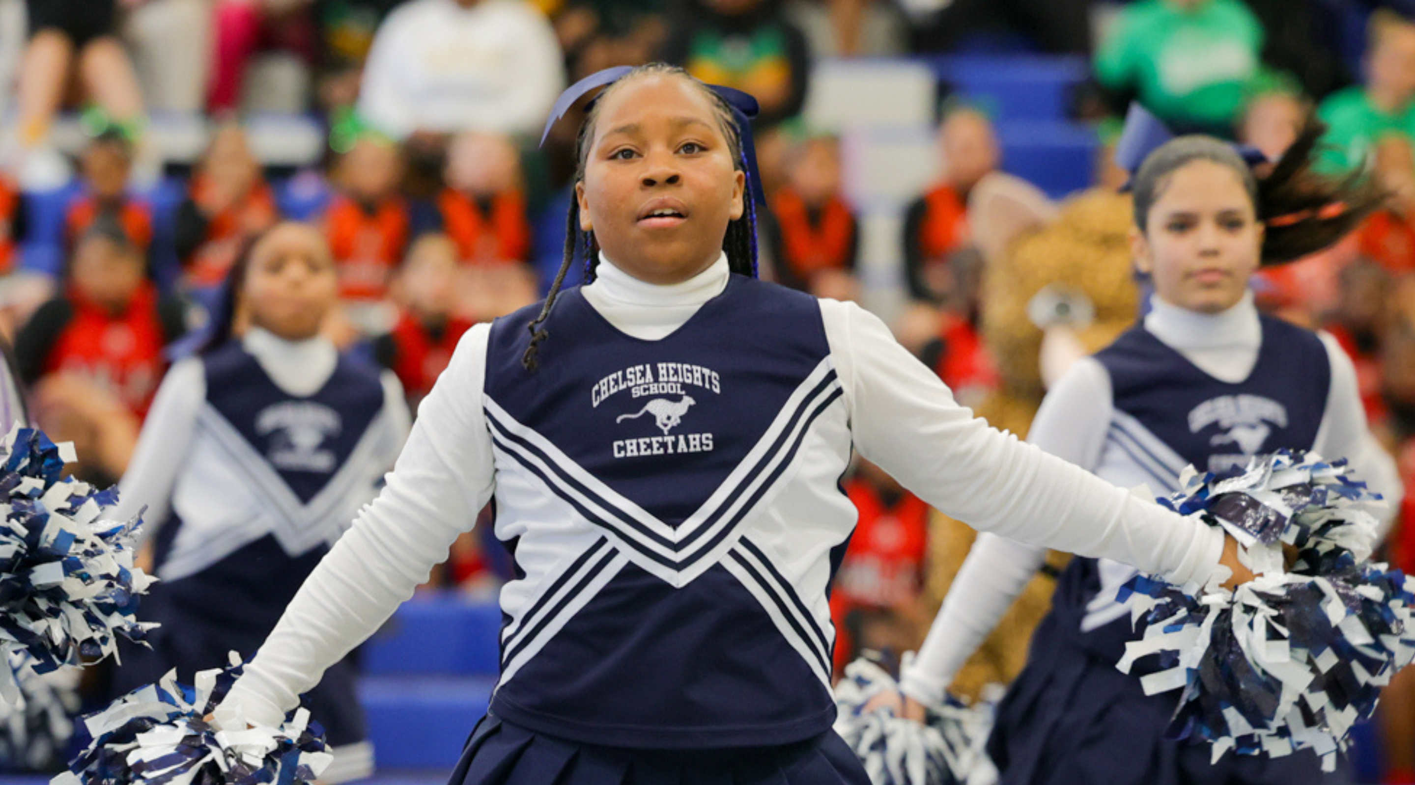 The image shows a group of cheerleaders in uniform, with one cheerleader in the foreground performing a routine while the others stand behind her.