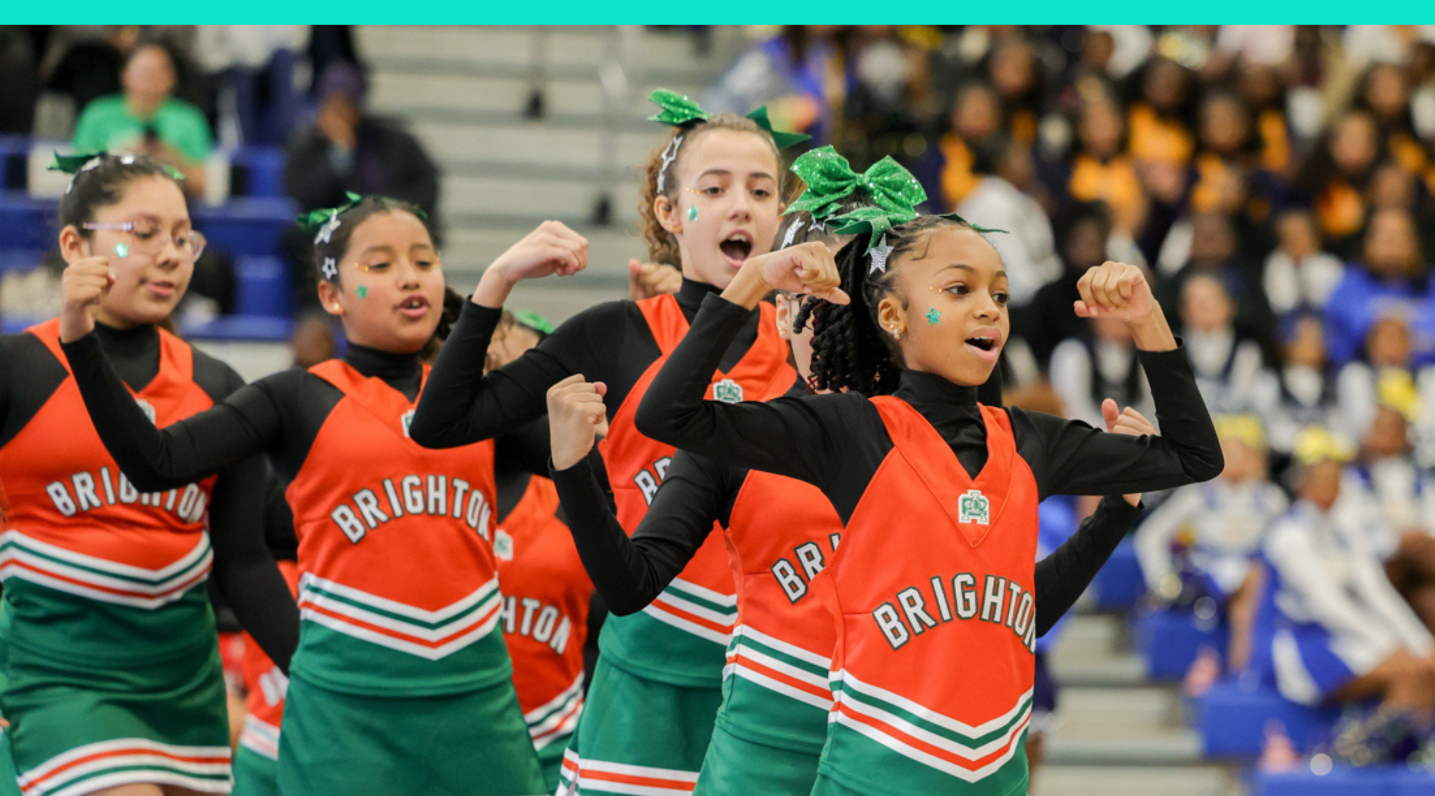 A group of cheerleaders in bright orange and green uniforms performing a synchronized routine on a stage with a colorful backdrop.