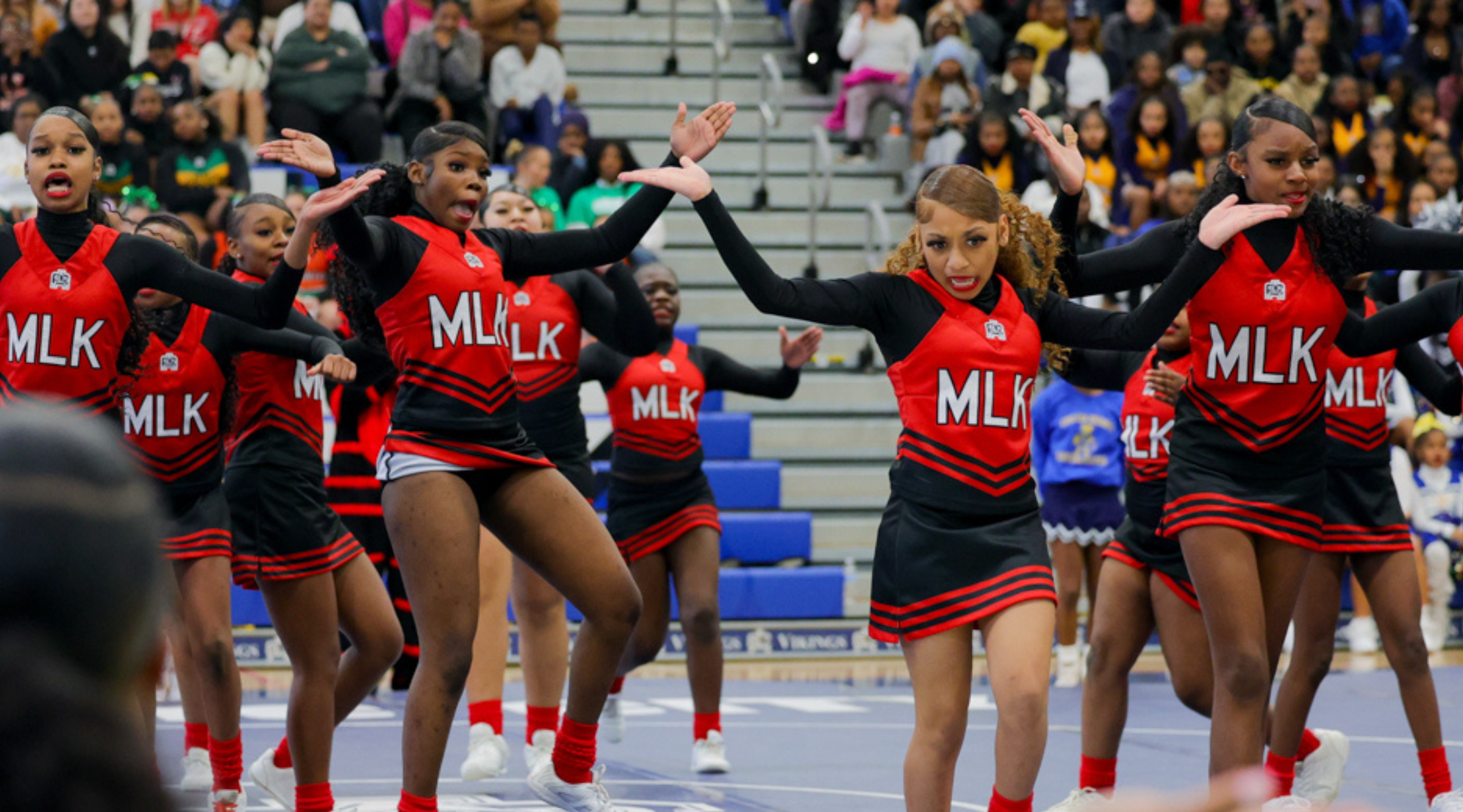 A group of cheerleaders in red and black uniforms with "MLK" printed on them are performing on a basketball court surrounded by a crowd of spectators.