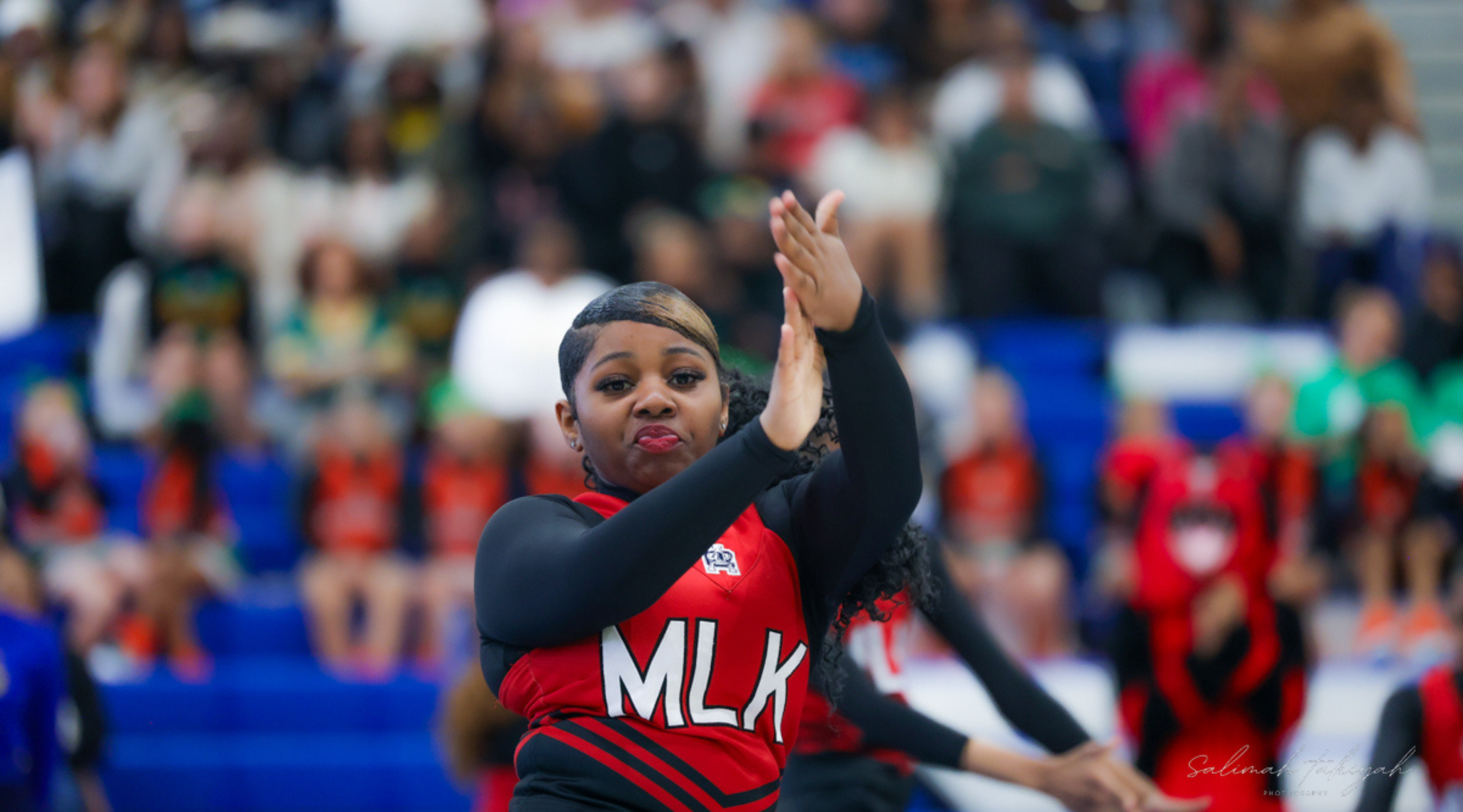 A person wearing a red jersey with "MLK" printed on it, standing in a crowd of people, with their arms raised.
