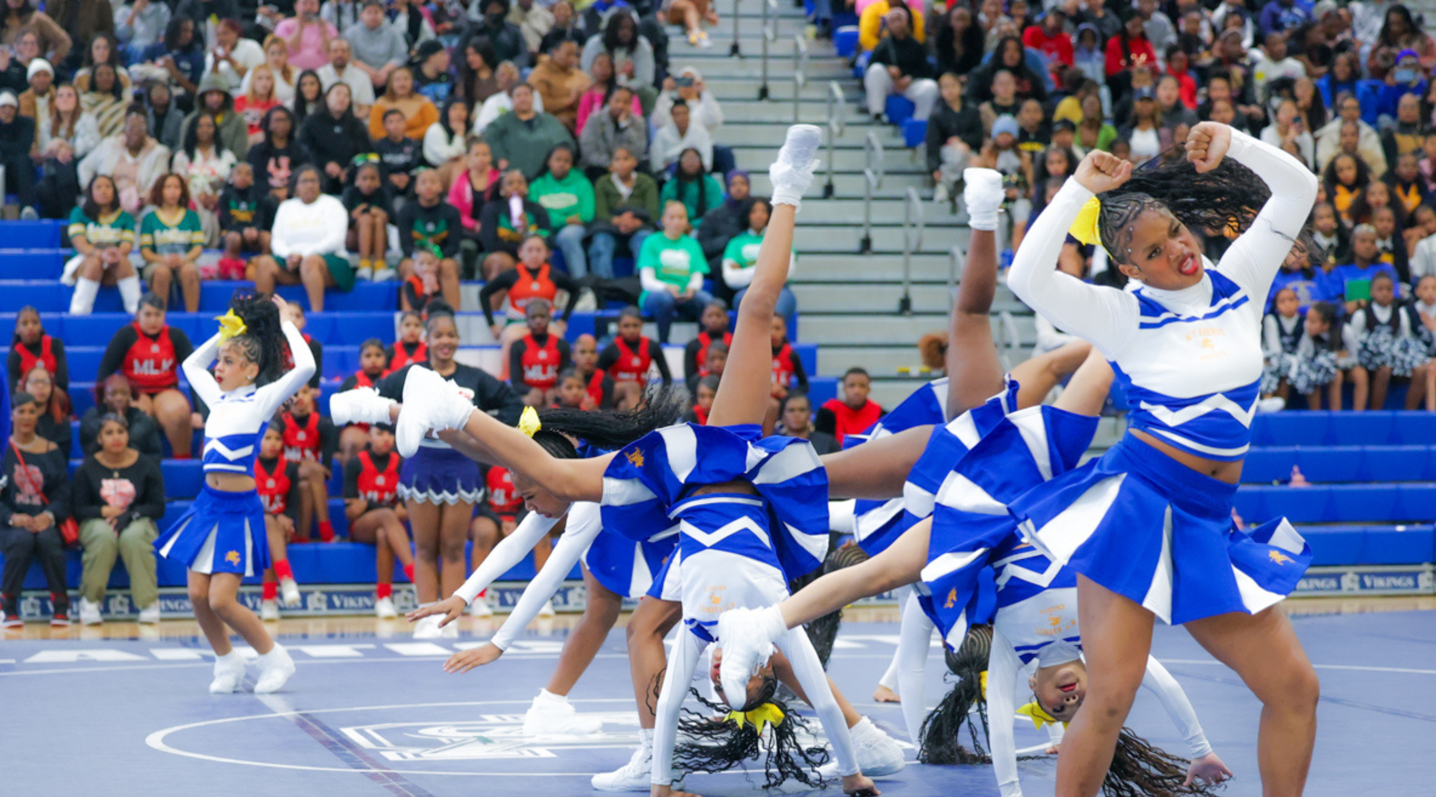 A group of cheerleaders in blue and white uniforms performing acrobatic stunts on a basketball court, with a large crowd of spectators visible in the background.