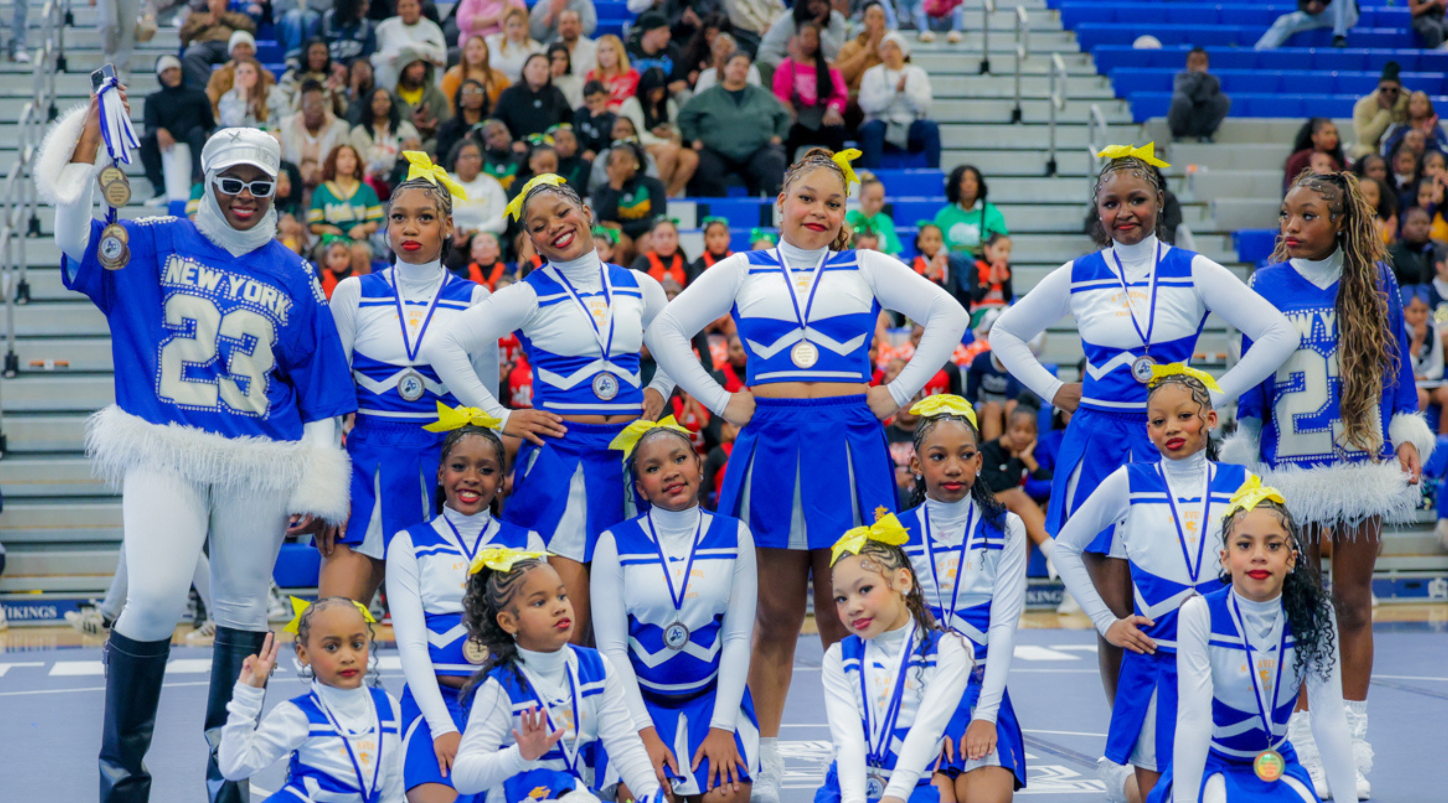 A group of cheerleaders in blue and white uniforms performing on a stadium stage, with a crowd of spectators visible in the background.