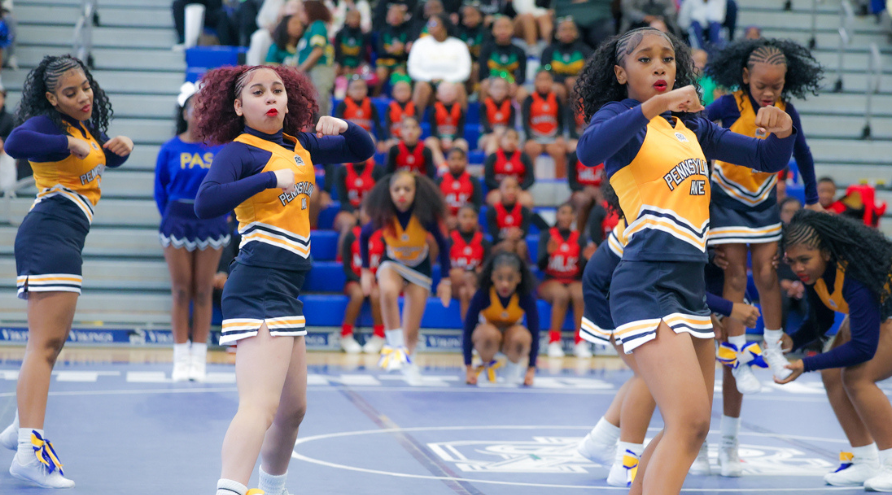 A group of cheerleaders in blue and yellow uniforms performing on a basketball court, with a crowd of spectators visible in the background.