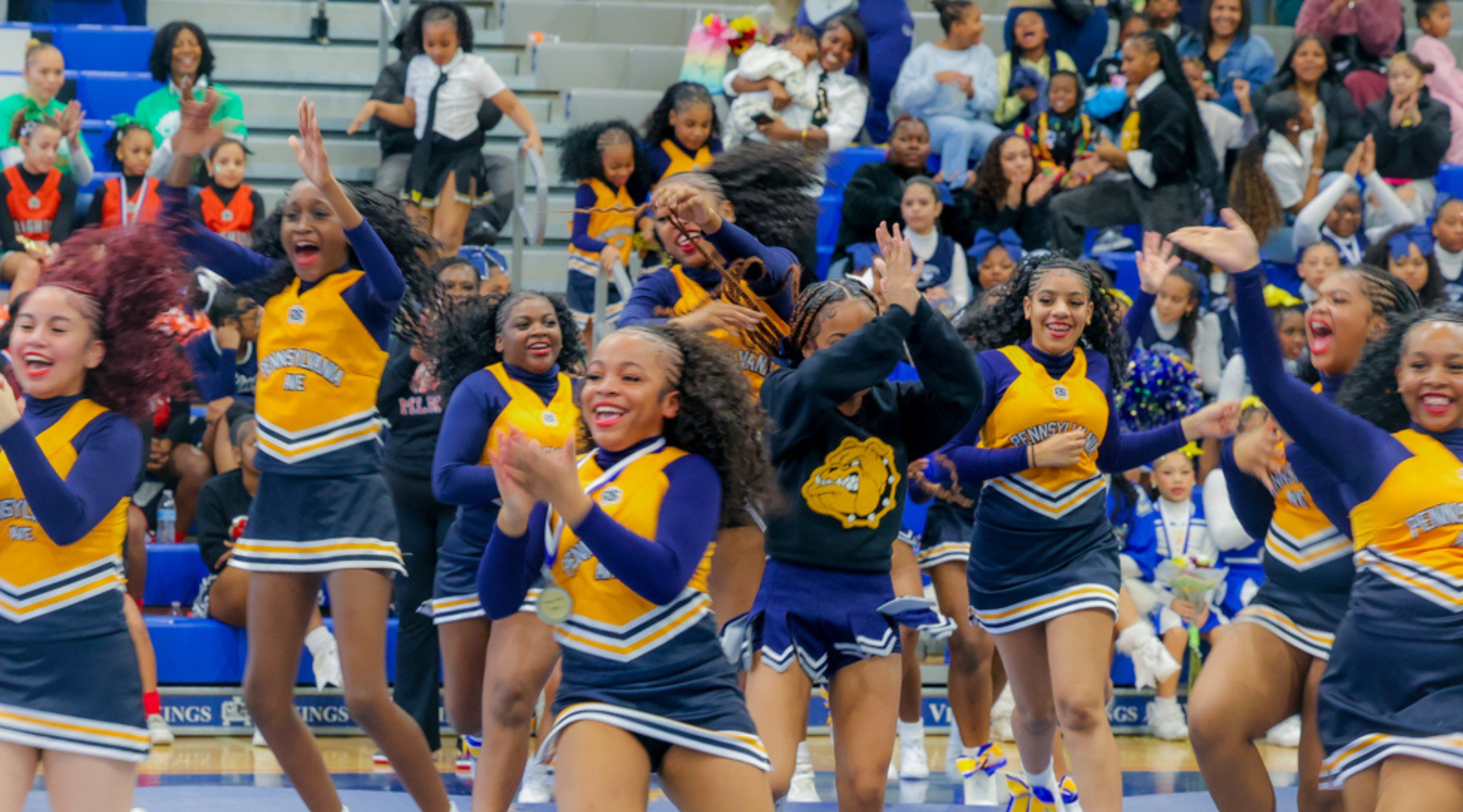A group of cheerleaders in colorful uniforms enthusiastically performing on a basketball court surrounded by spectators.