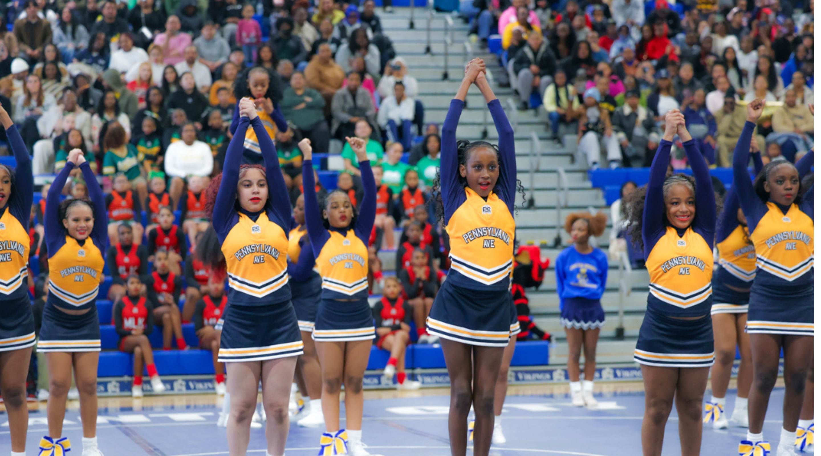 A large group of cheerleaders in colorful uniforms stand on a stadium field, surrounded by a crowd of spectators in the background.