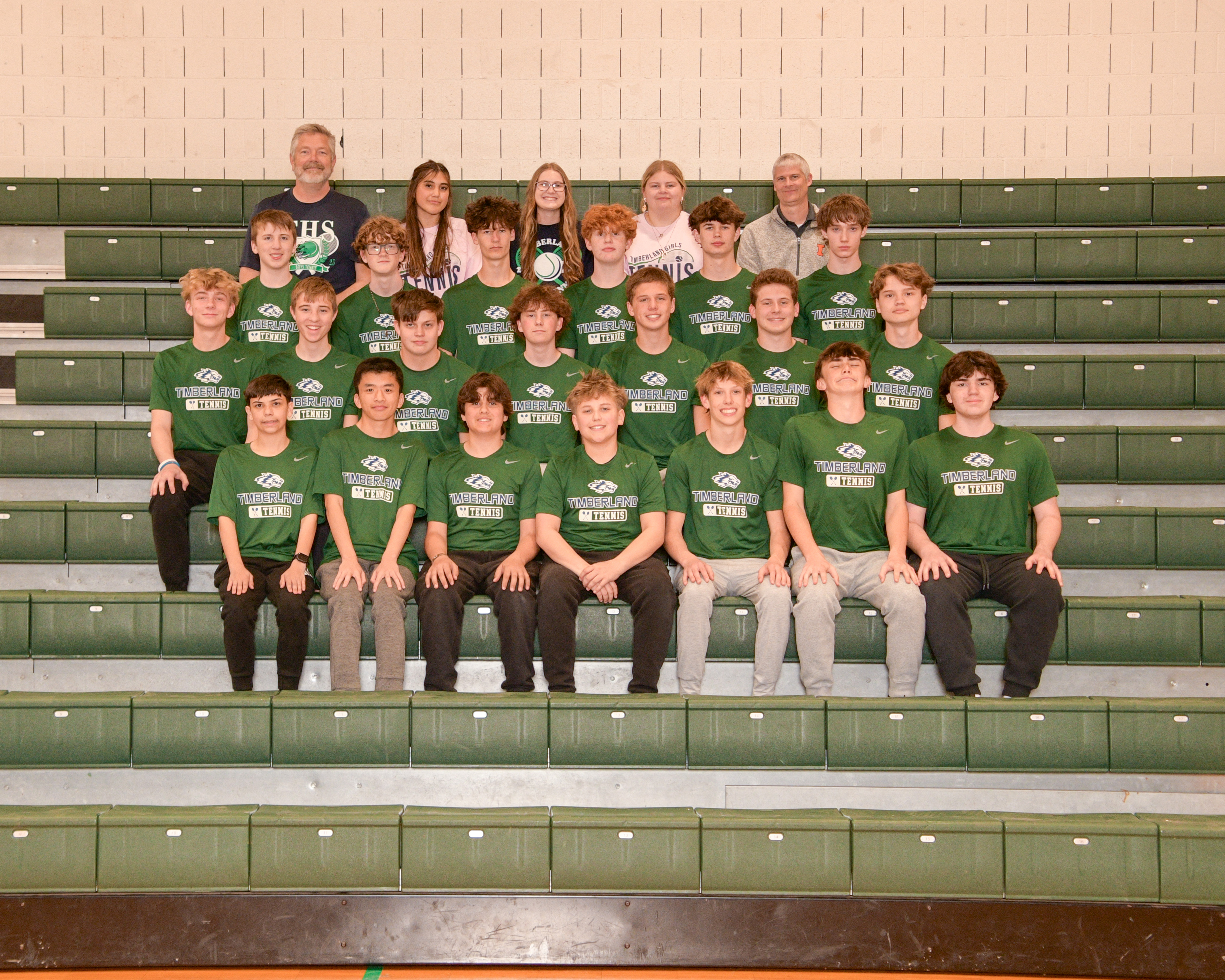 A junior varsity tennis team, posing together on stadium seating with green uniforms and a logo visible.